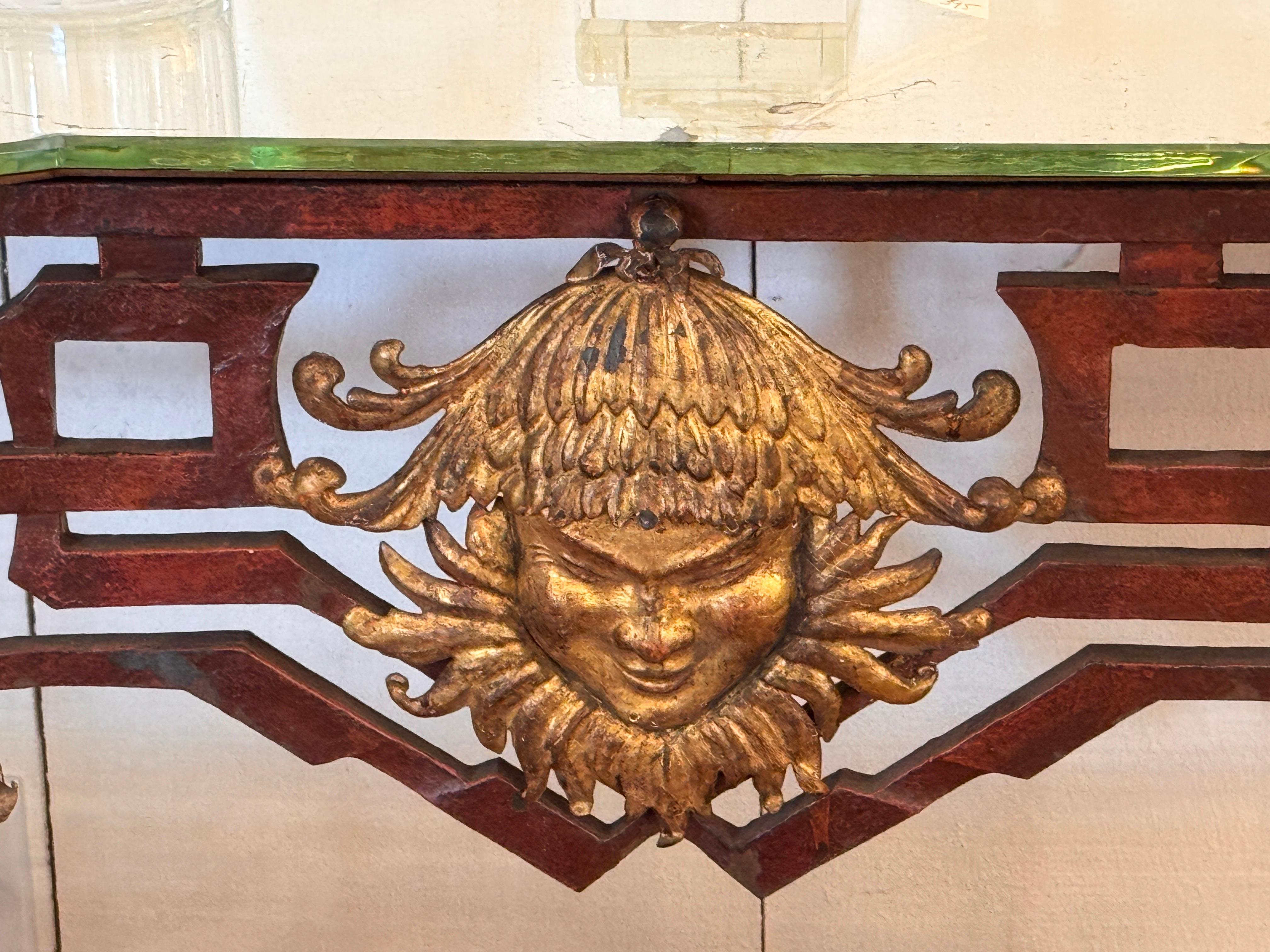 A striking Italian Deco console table featuring a long mirrored top supported by a curvaceous iron base. The design incorporates a geometric fretwork frieze, flared legs, and gilt foliate drop details. At center, a bold gilt mask medallion anchors
