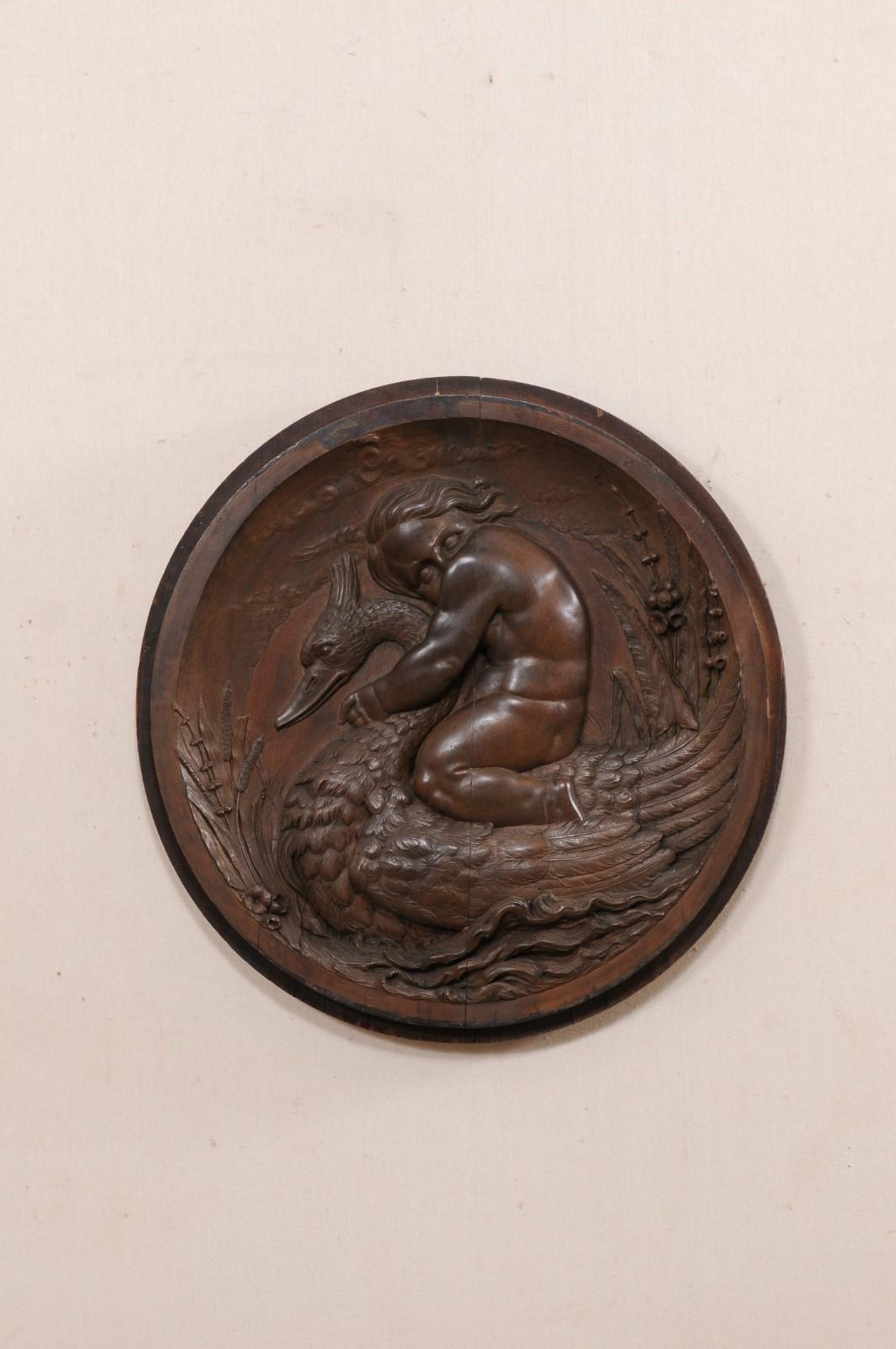 An Italian carved-wood hanging wall plaque from the early 19th century. This antique wall decoration from Italy is round in shape and features a hand-carving depiction of a putto hanging onto lovingly, as if riding, atop a water fowl. Waving water