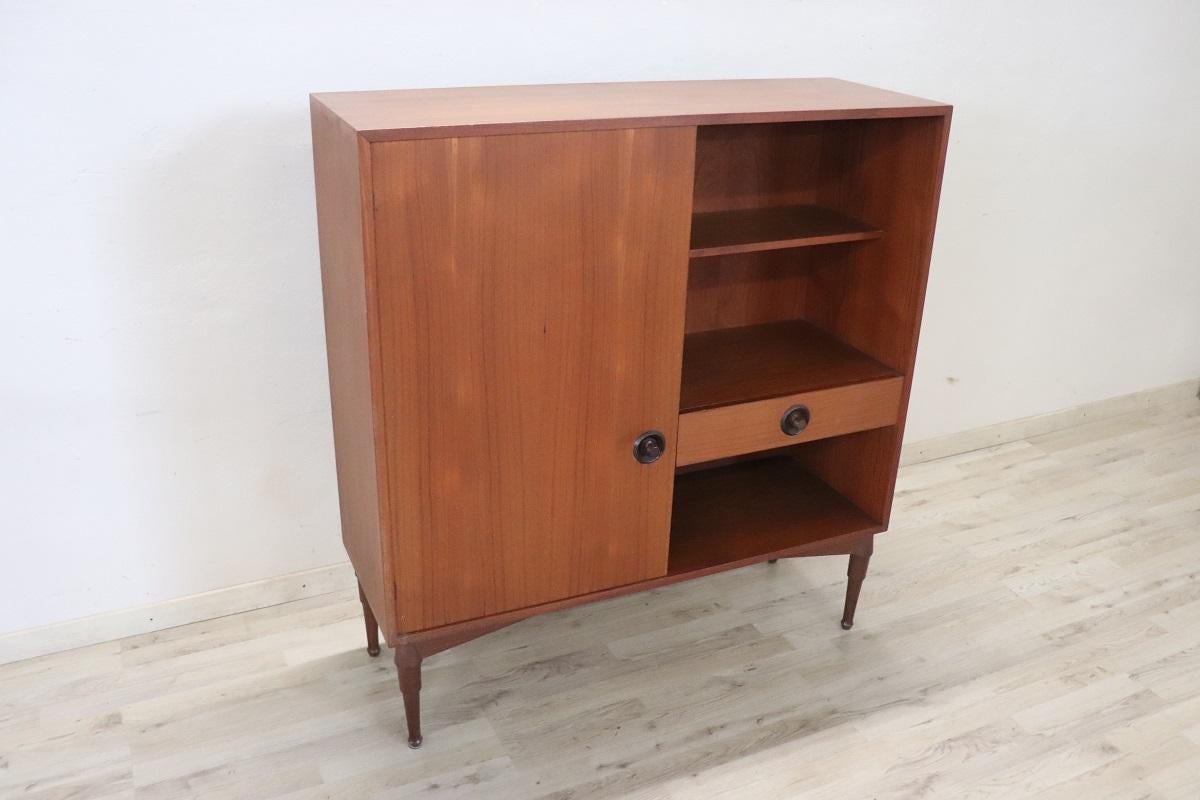 Beautiful 1960s Italian design highboard in teak veneer. Characterized by a very linear minimalist style. Equipped with one side with exposed shelves and a drawer, while the other side with a door and two internal shelves. A piece of furniture that