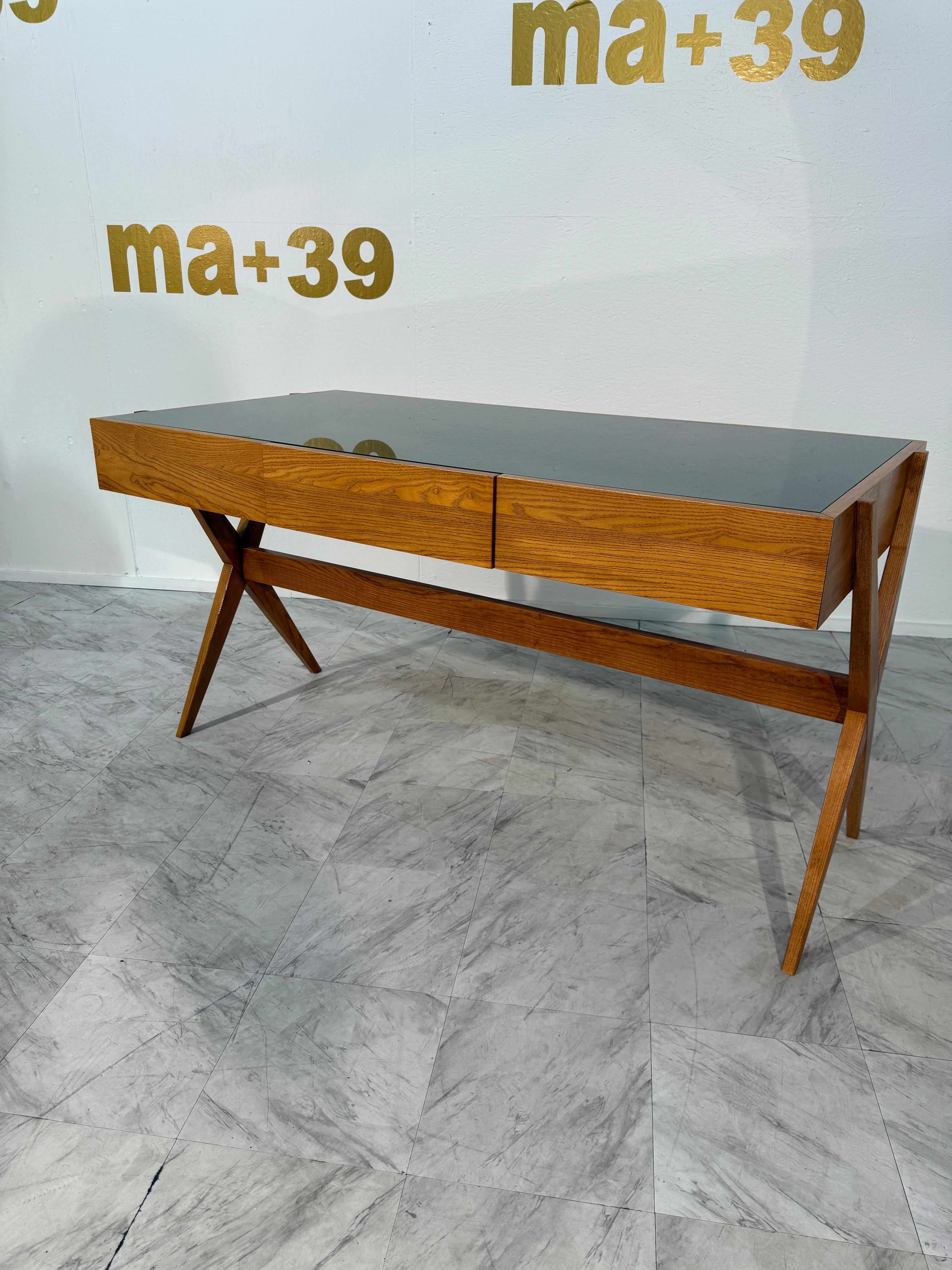 Se trata de un escritorio italiano moderno de alrededor del año 2000.
Tiene una cálida estructura de madera con patas en ángulo y una tapa de cristal verde oscuro.
El frontal presenta dos cajones delgados sin tiradores, manteniendo el aspecto limpio