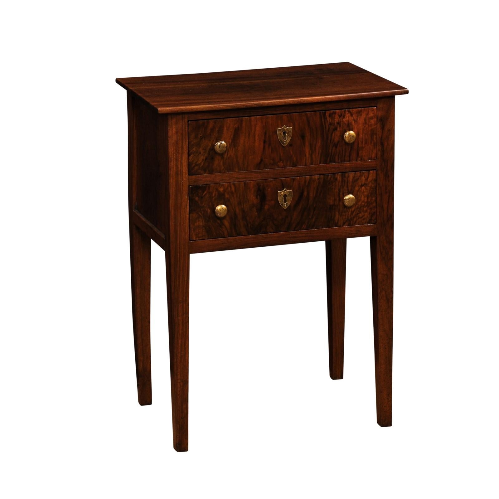 An Italian Directoire period walnut bedside table from the 19th century, with two drawers, butterfly veneer on the façade, brass hardware, shield shaped escutcheons and tapered legs. This Italian Directoire period walnut bedside table, dating back