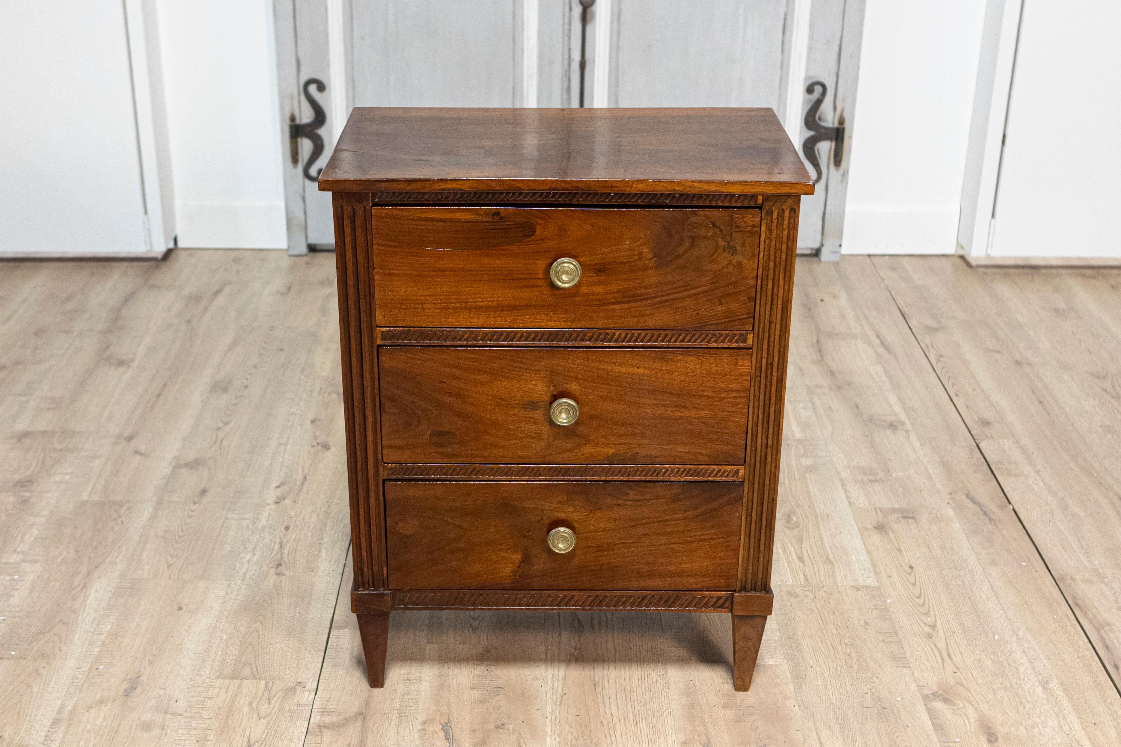 An Italian Directoire period bedside chest from the 18th century, in solid walnut, with three drawers, carved accents and fluted side posts. This exquisite Italian Directoire period bedside chest from the 18th century showcases the elegance and