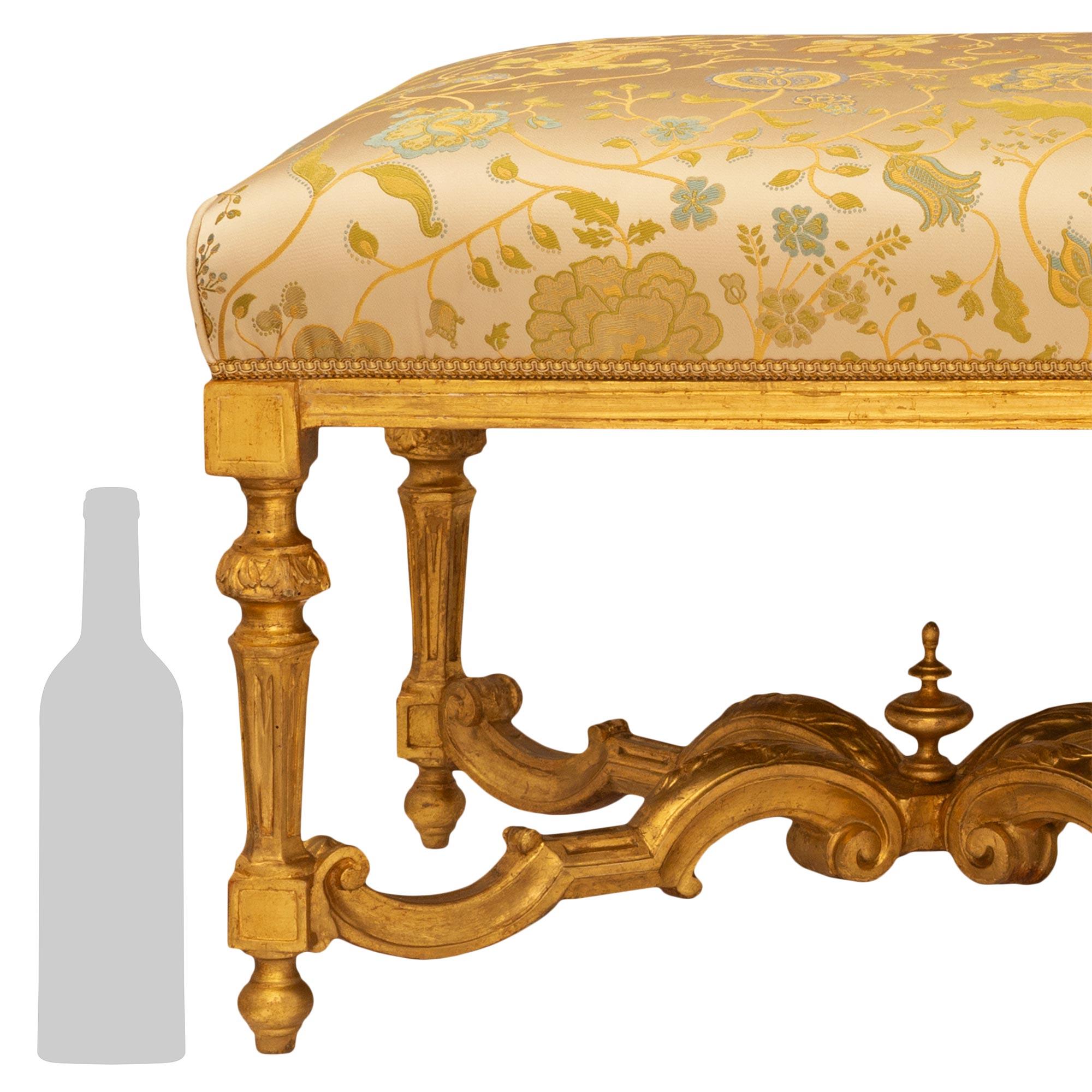 An elegant Italian early 18th century Louis XIV period giltwood bench. The long bench is raised on six legs with topie shape feet below a circular fluted tapered column, foliate decorated ball, and square block above. The legs are joined by an X