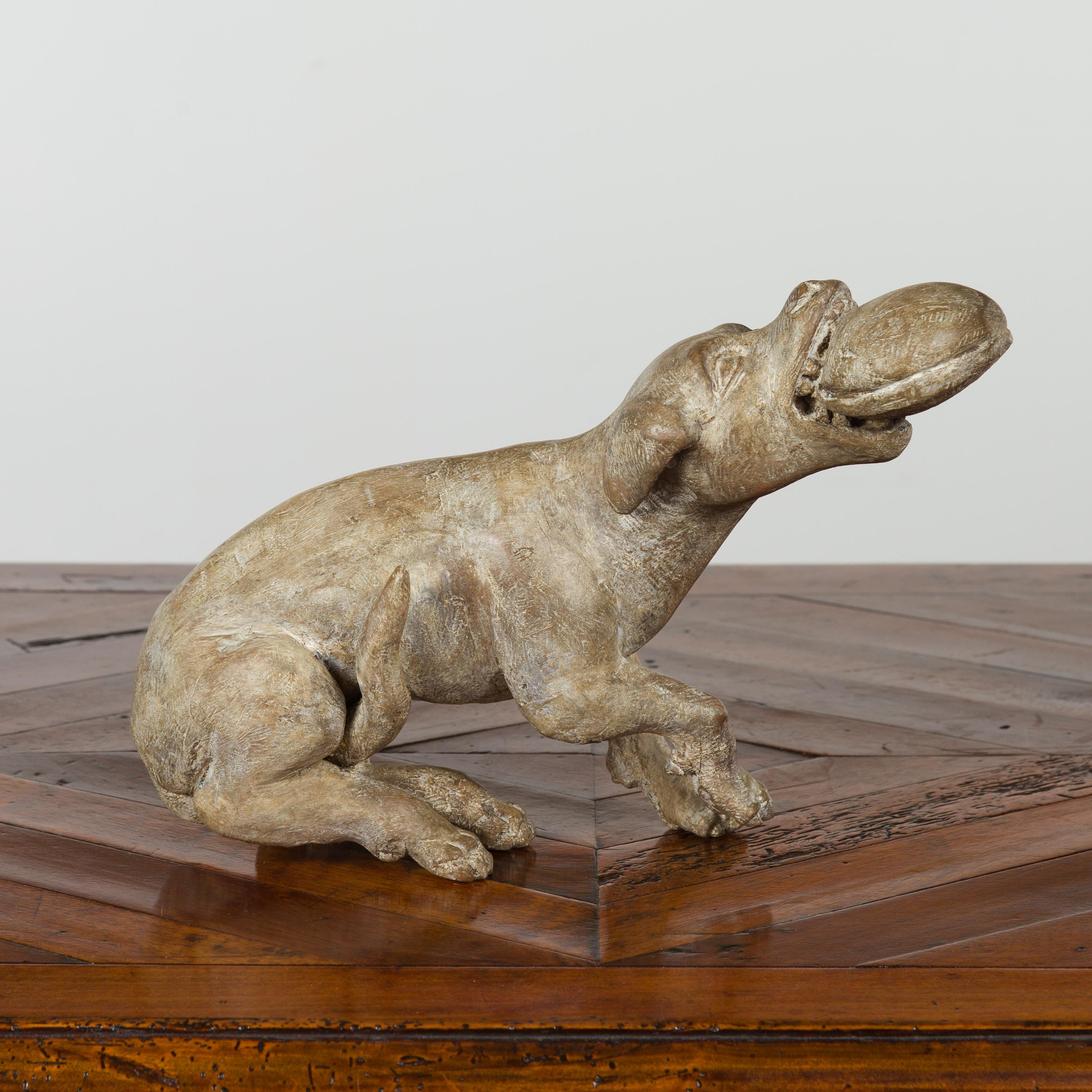 An Italian early 19th century carved wooden dog playing with his toy. Hand carved in Italy during the early years of the 19th century, this wooden dog charms us with his playful attitude. Sitting on his behind, he is holding in his mouth his toy, as