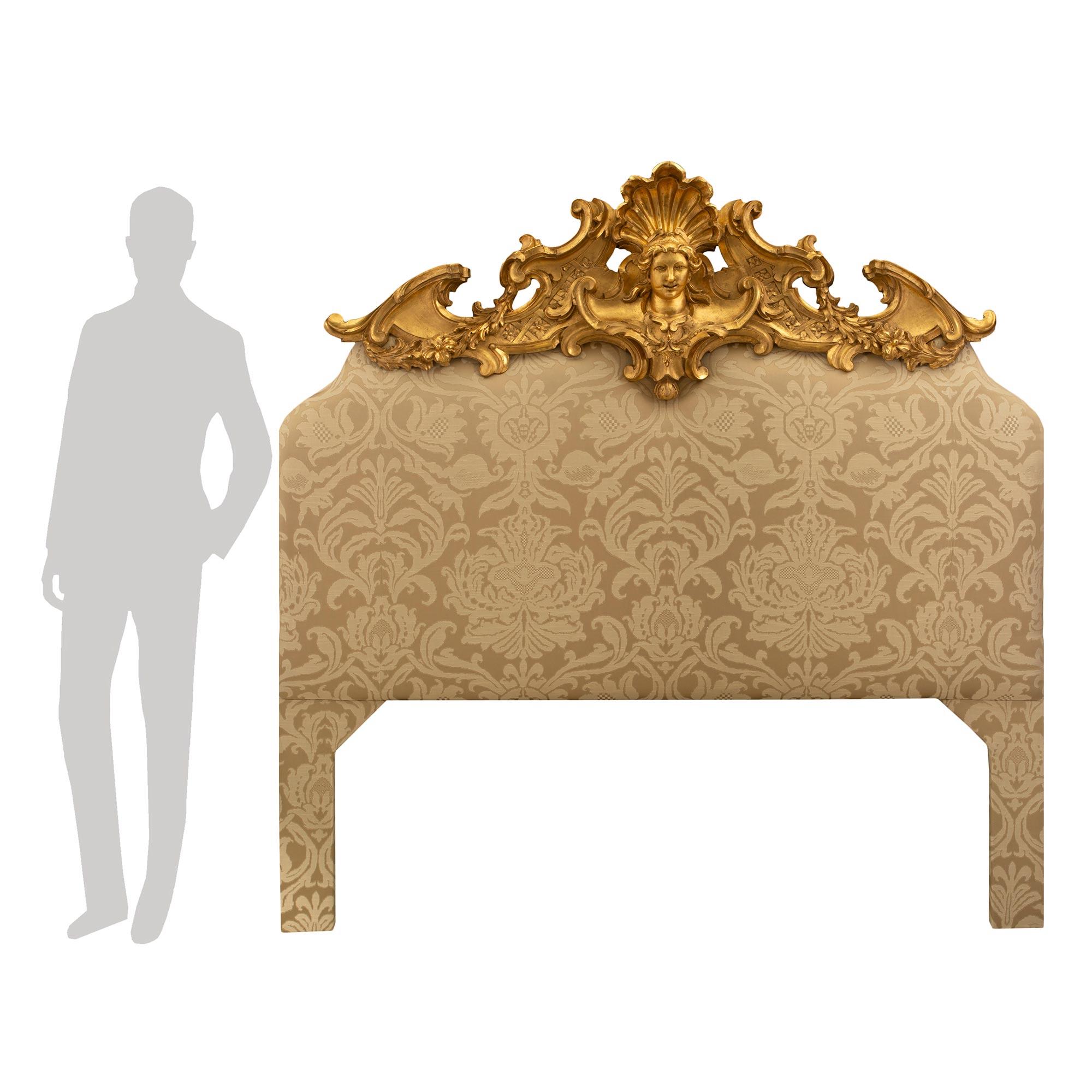 A striking Italian early 19th century Louis XV st. giltwood upholstered headboard. The headboard displays an impressive and most decorative carved giltwood top crown. The top crown is centered by a richly carved maiden's face with a beautiful