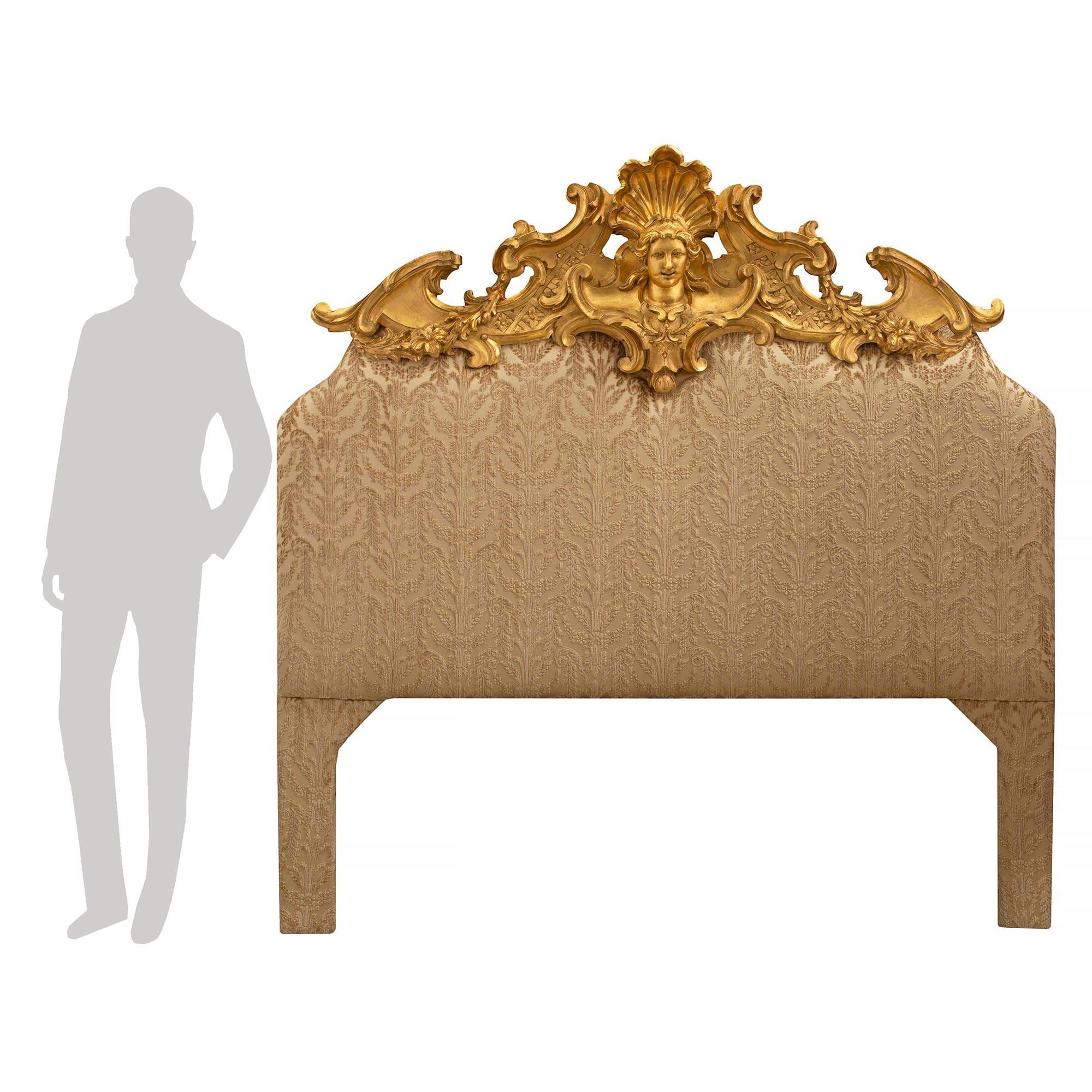 A striking Italian early 19th century Louis XV st. upholstered and giltwood headboard. The headboard displays an impressive and most decorative carved giltwood top crown. The top crown is centered by a richly carved maiden's face with a beautiful
