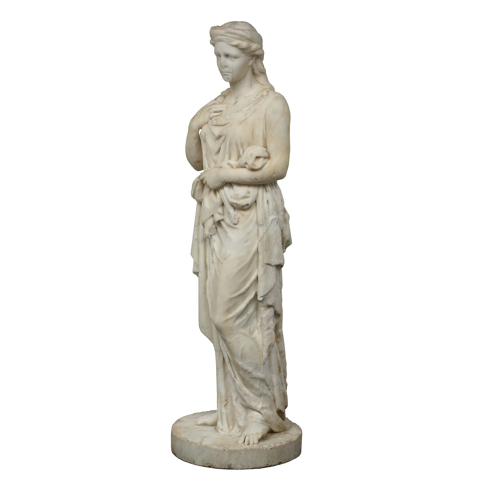 A lovely Italian early 19th century solid white Carrara marble statue of a young maiden. The seasoned figure is raised on a solid circular base. The bare footed maiden is draped in classical dress, one hand gathering up her skirt and her other hand