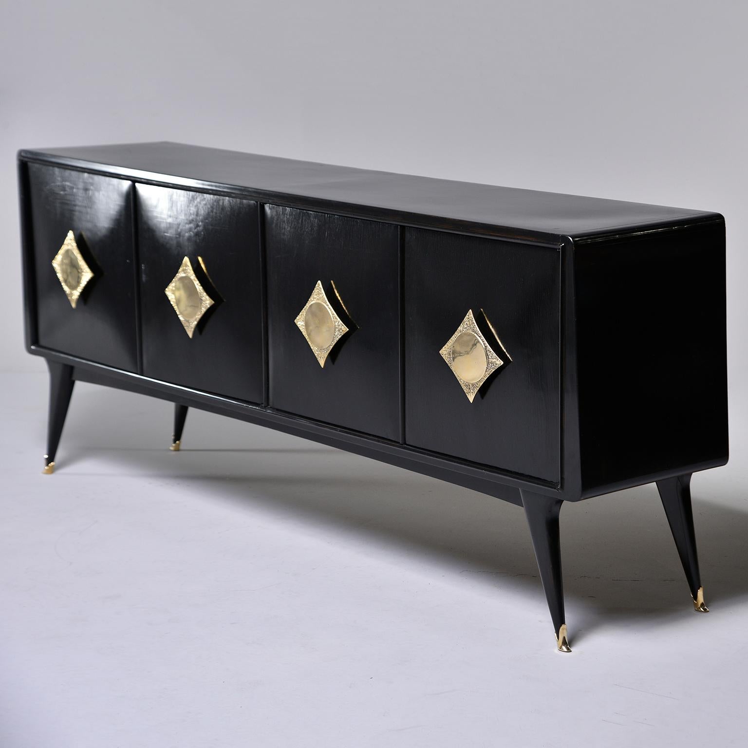Circa 1950s Italian sideboard has an ebonised finish, angled tapered legs with brass sabots, two storage compartments with internal shelf and four hinged doors with oversized decorative brass knobs. Unknown maker.