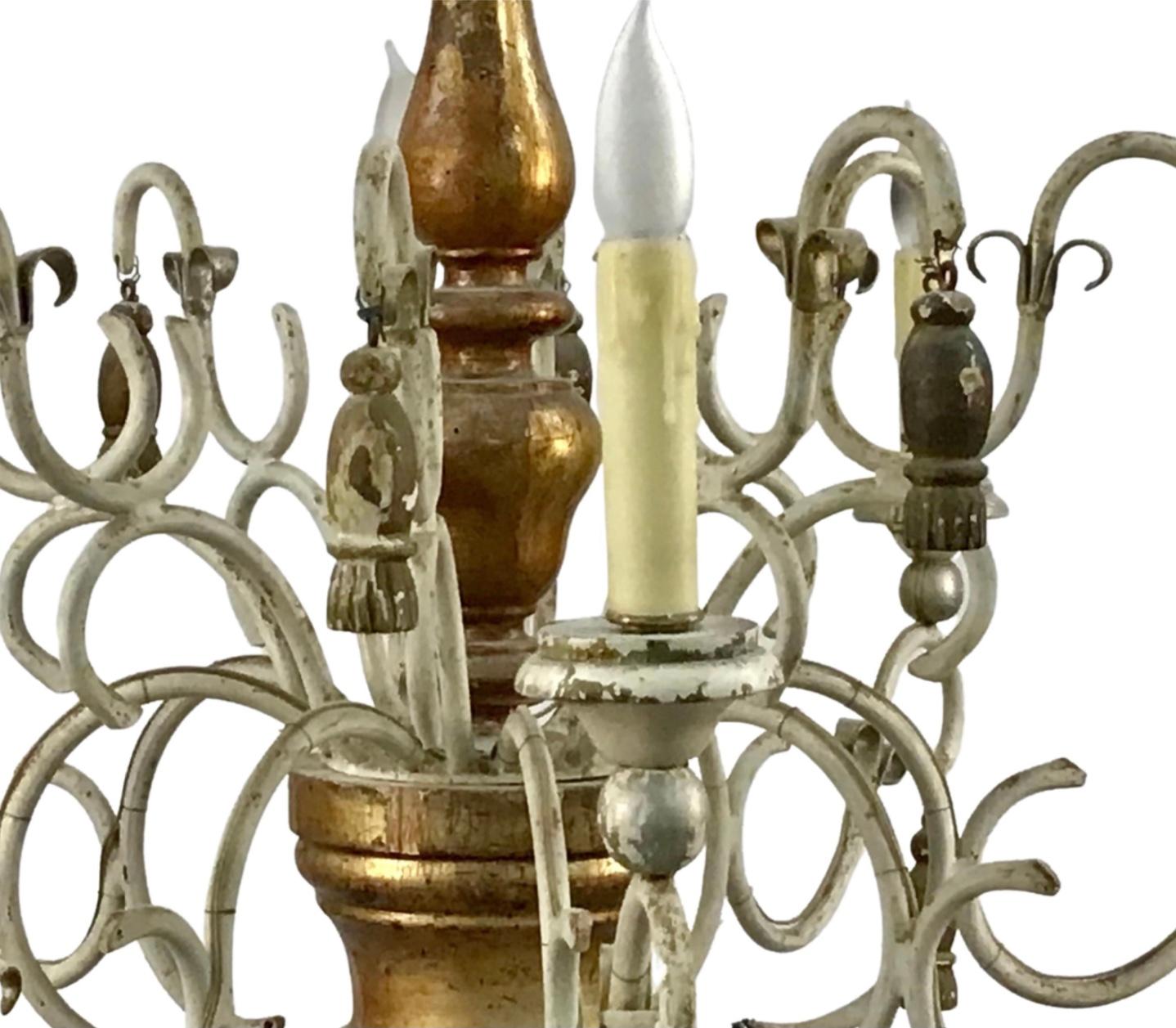 An antique Italian eight arm chandelier. Aged patina and neutral colors with gold and silver gilt center wood section. Scrolling wrought iron arms with carved and turned grey painted wood baluster, gilt wood tassel and bell hanging decoration, all