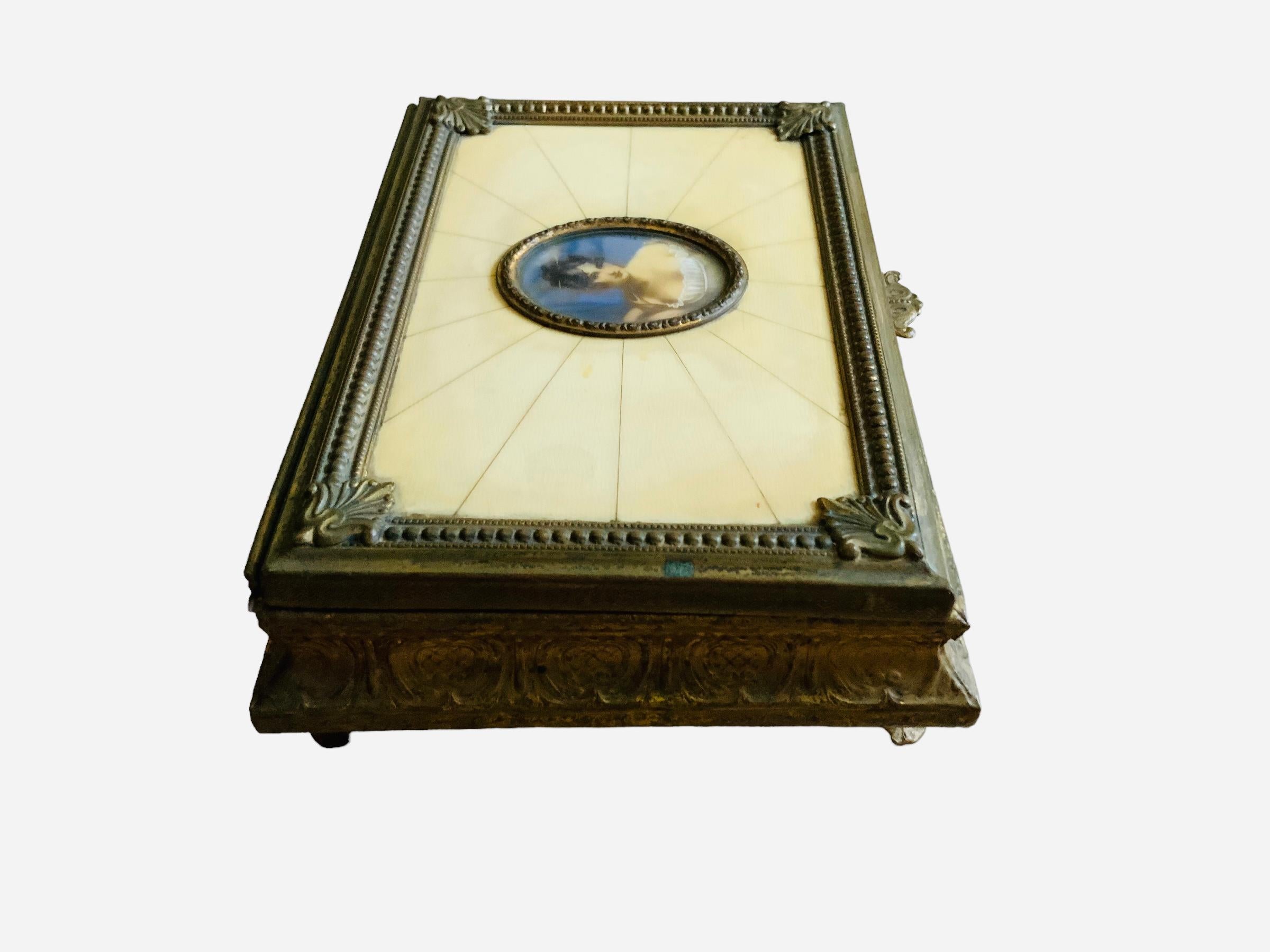 This is an Italian bronzed metal square hinged lidded music box. It depicts a lid decorated in the center with a 19th century hand painted portrait of Madame Recamier adorned with a patinated metal oval frame. A faux ivory panel around enhances it.