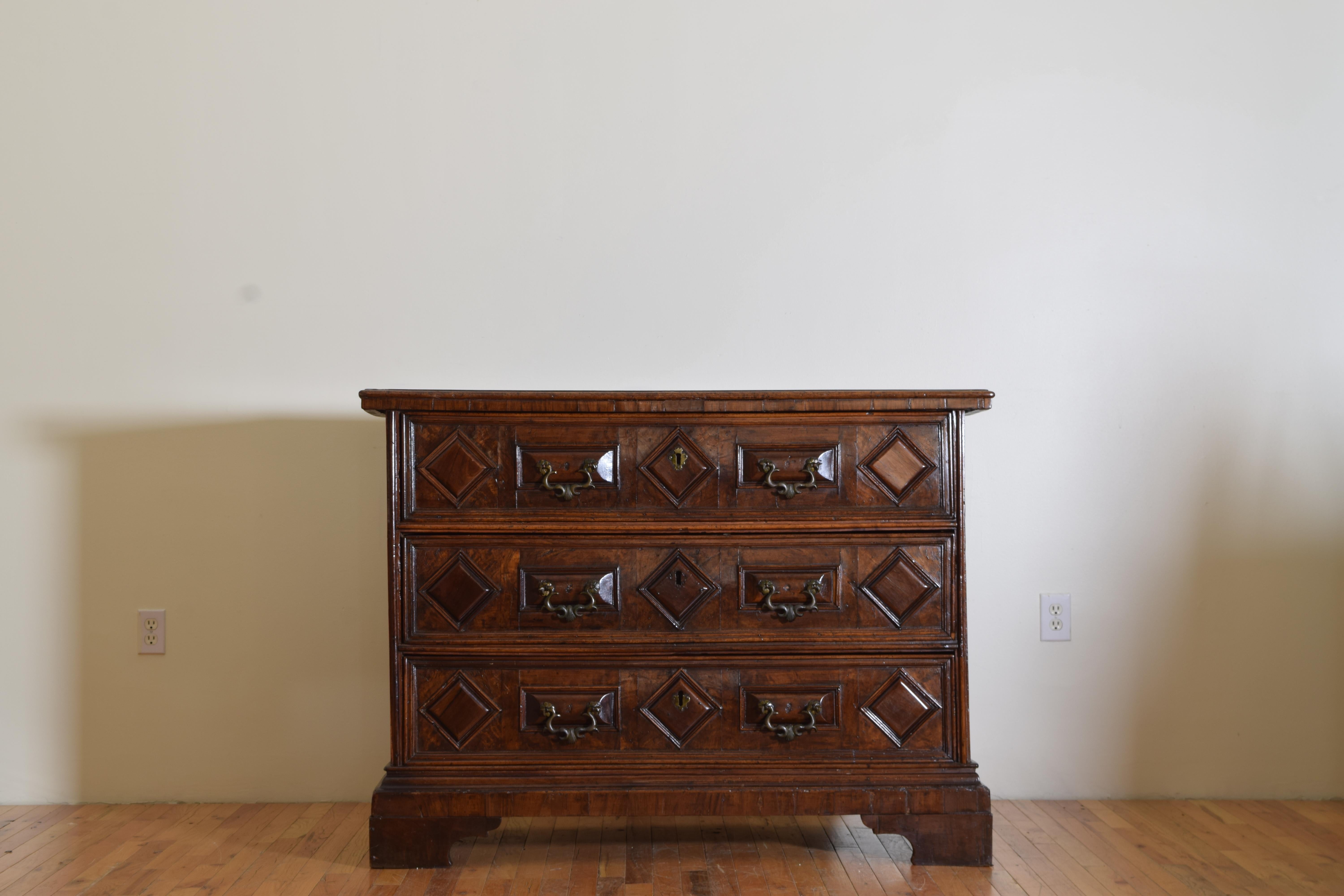 having a rectangular top with slightly overhanging apron above a conforming case housing three drawers each with carved raised panels of lozenges and rounded rectangles with bronze handles, the case raised on shaped bracket feet.