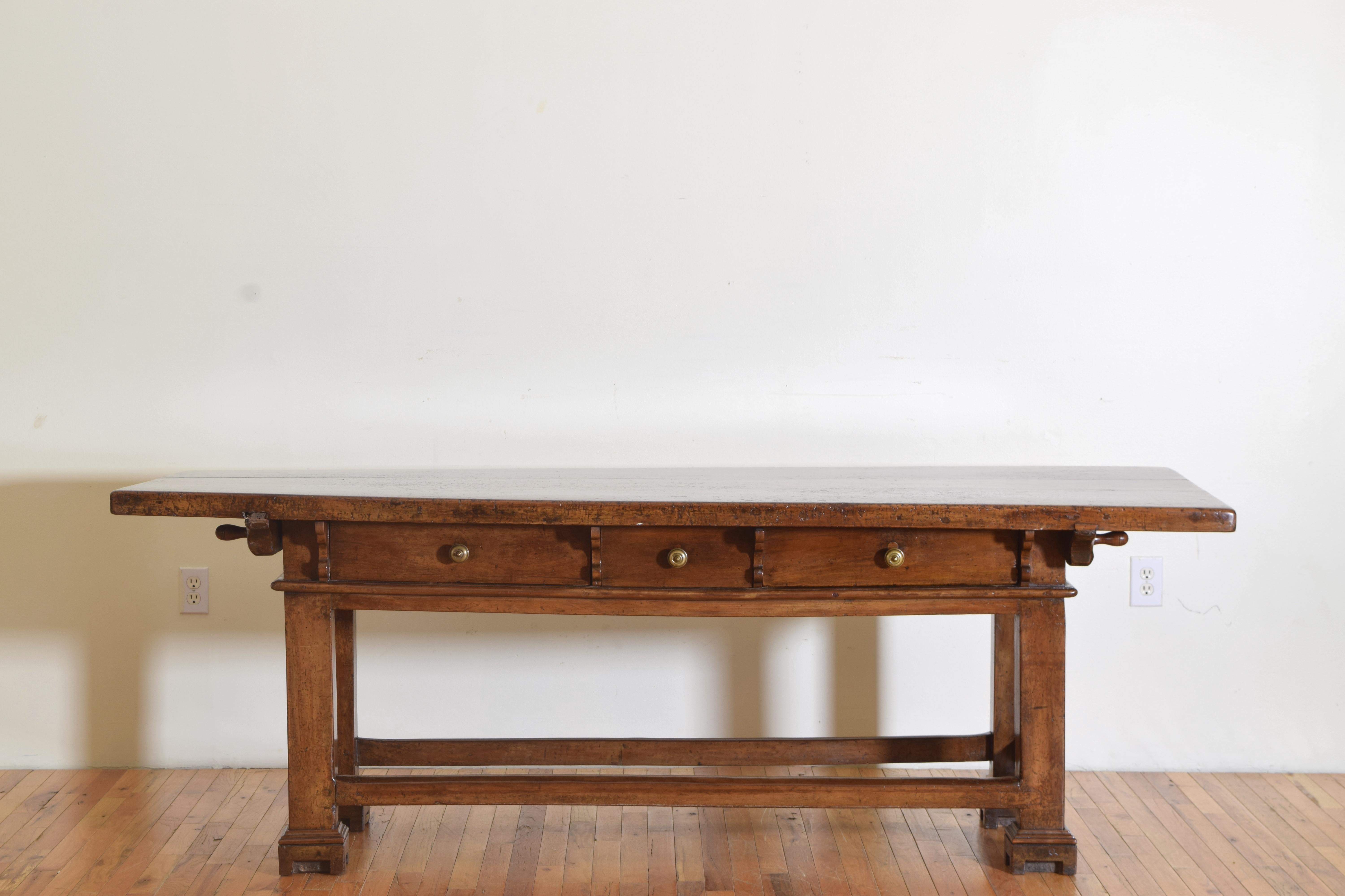 Having a thick walnut top resting on a case housing three drawers, the top and case connected by turned wooden pegs, the drawers separated by decorative pearwood buttresses and retaining antique bronzes, with a thick decorative molding at the bottom