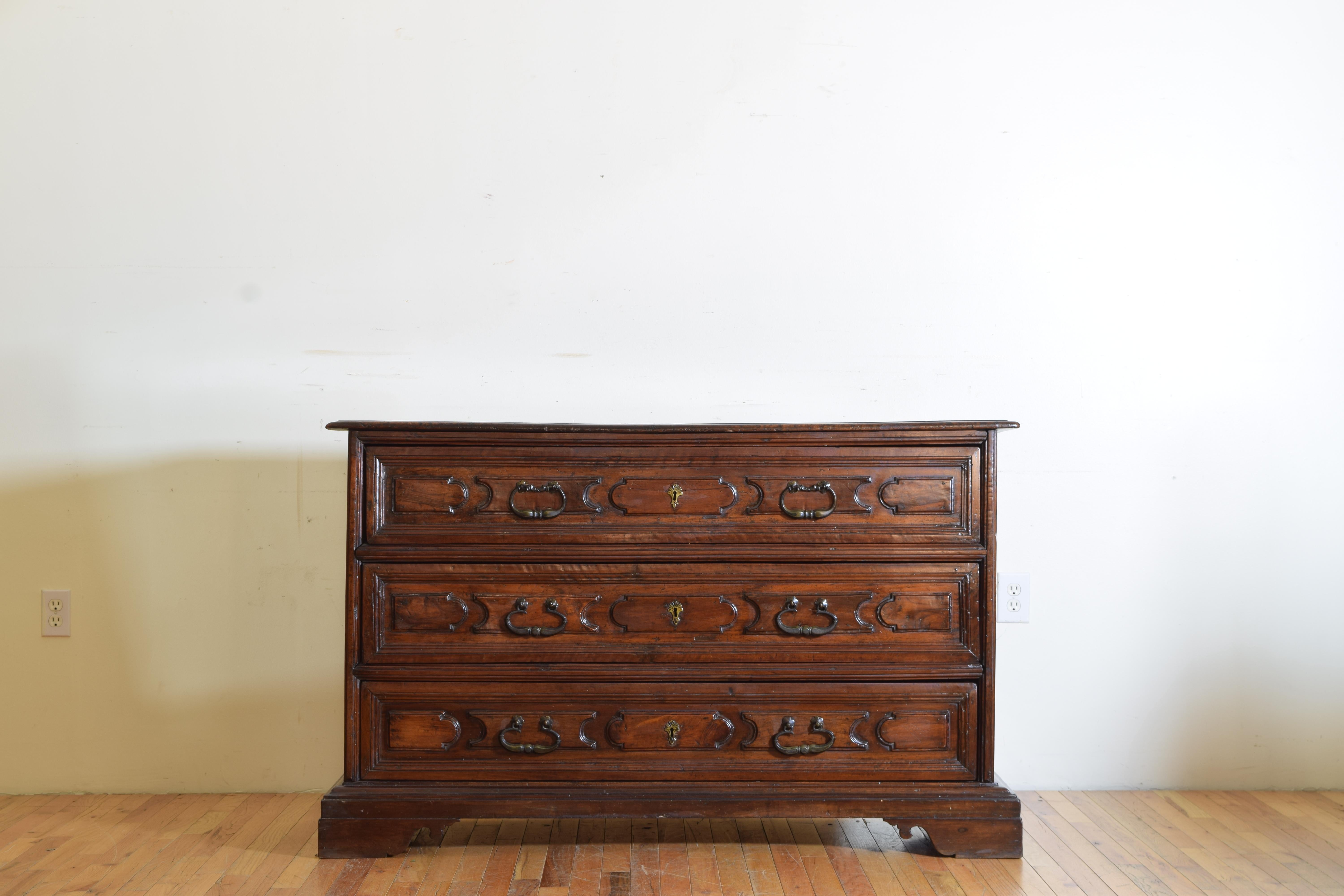 The rectangular top with a molded edge above a conforming case housing three paneled drawers with original patinated brass pulls, the sides of the case with large raised panels, the whole resting on a plinth form base atop shaped bracket feet, 17th