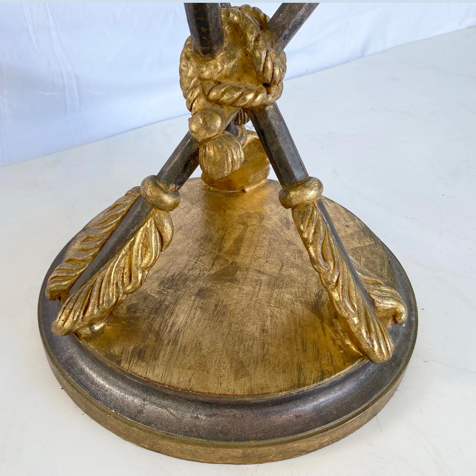 Accent table, in Empire taste, having a silver and gold gilt finish, with black accents; its round top of glass, on an apron stepped from the inside edge, raised on a trio of hand-carved, crossed arrows, bound by a tasseled cord, on a graduated