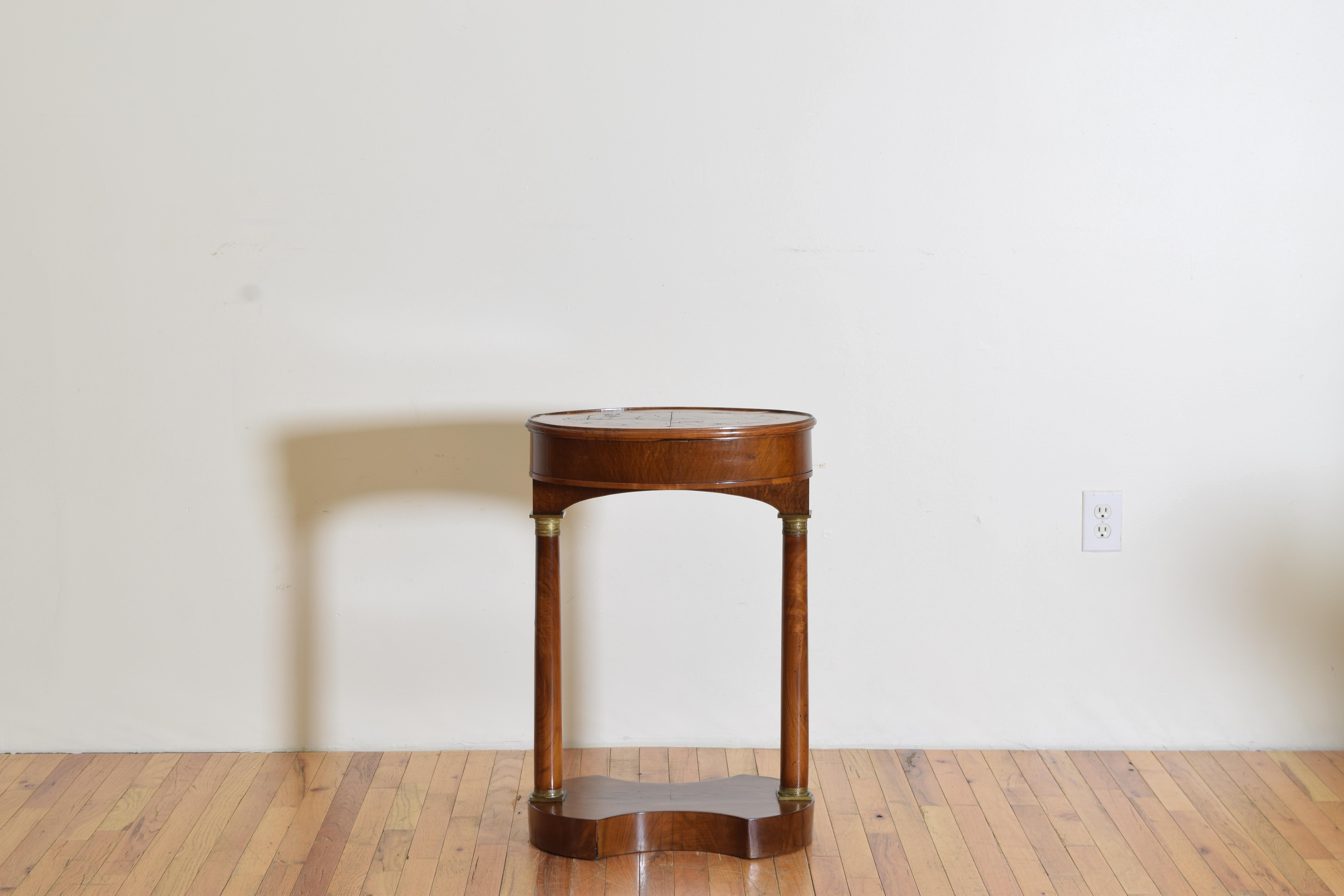 The elliptical shaped top with mixed veneers and a raised edge, the reverse of the top embossed in red leather, the top above a storage well and raised on columnar supports with brass mounted capitals resting atop a shaped base.