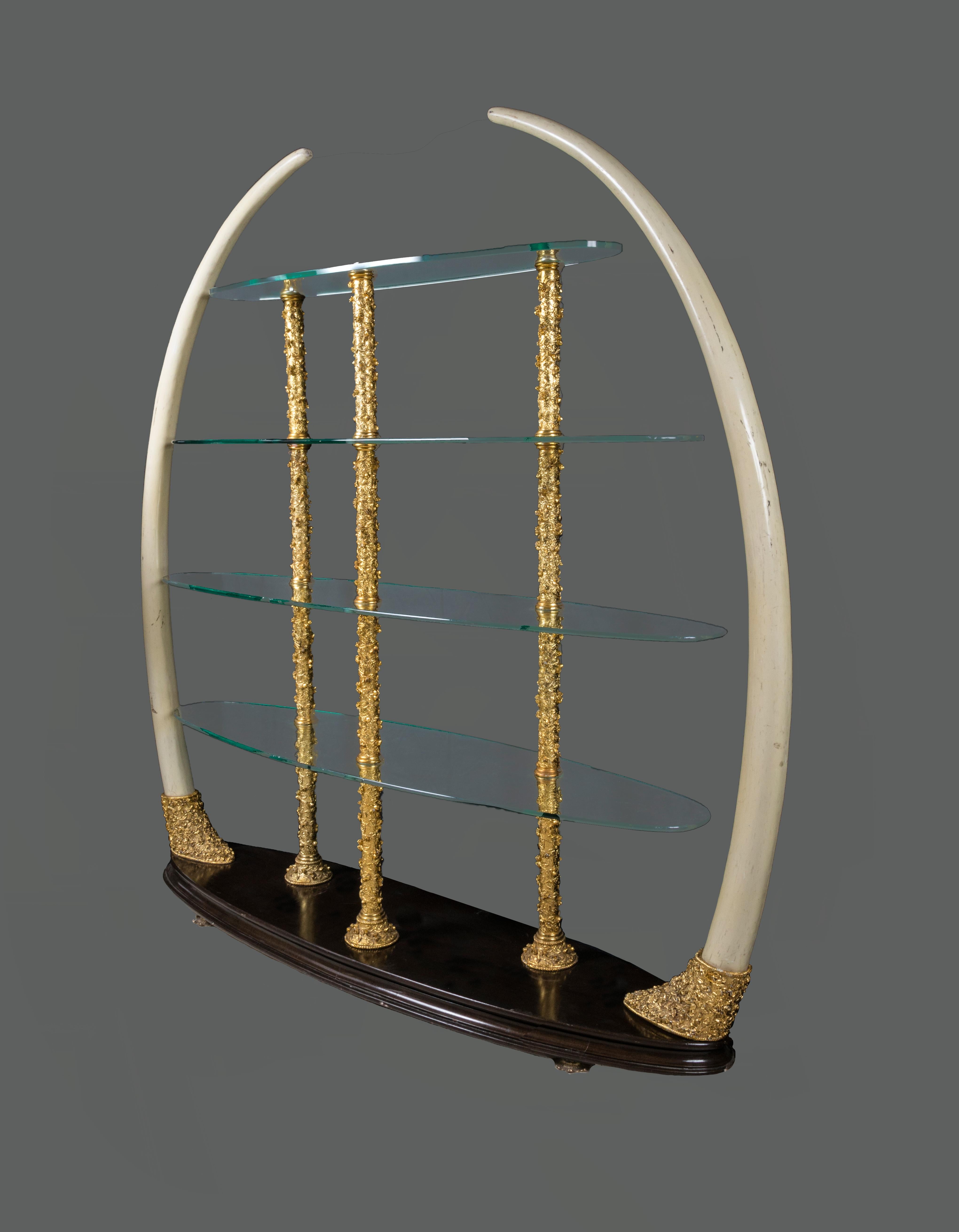 Very unique Italian vintage faux elephant tusk étagère, bronze structure studded with small golden flowers, four oval different sizes crystal shelves, oval wood base, brass flower feet. The Elephant tusks are made of resin, they look very realistic