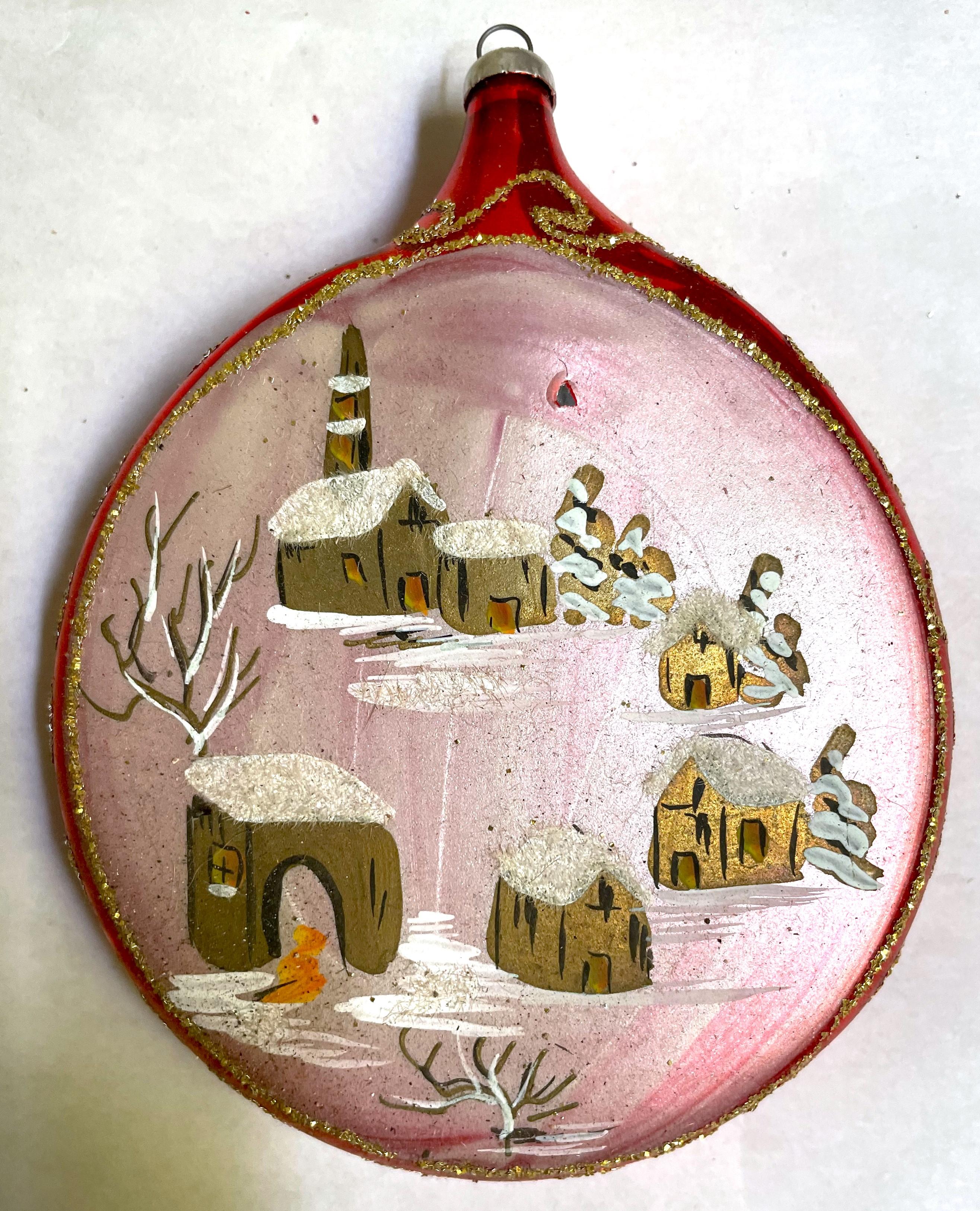 What a fantastic large shiny red mercury glass flattened ball Christmas tree ornament with a sweet snow covered village, including a bridge, hand painted on the front and swirls of gold glitter adorning the sides. It would be wonderful an a tree or