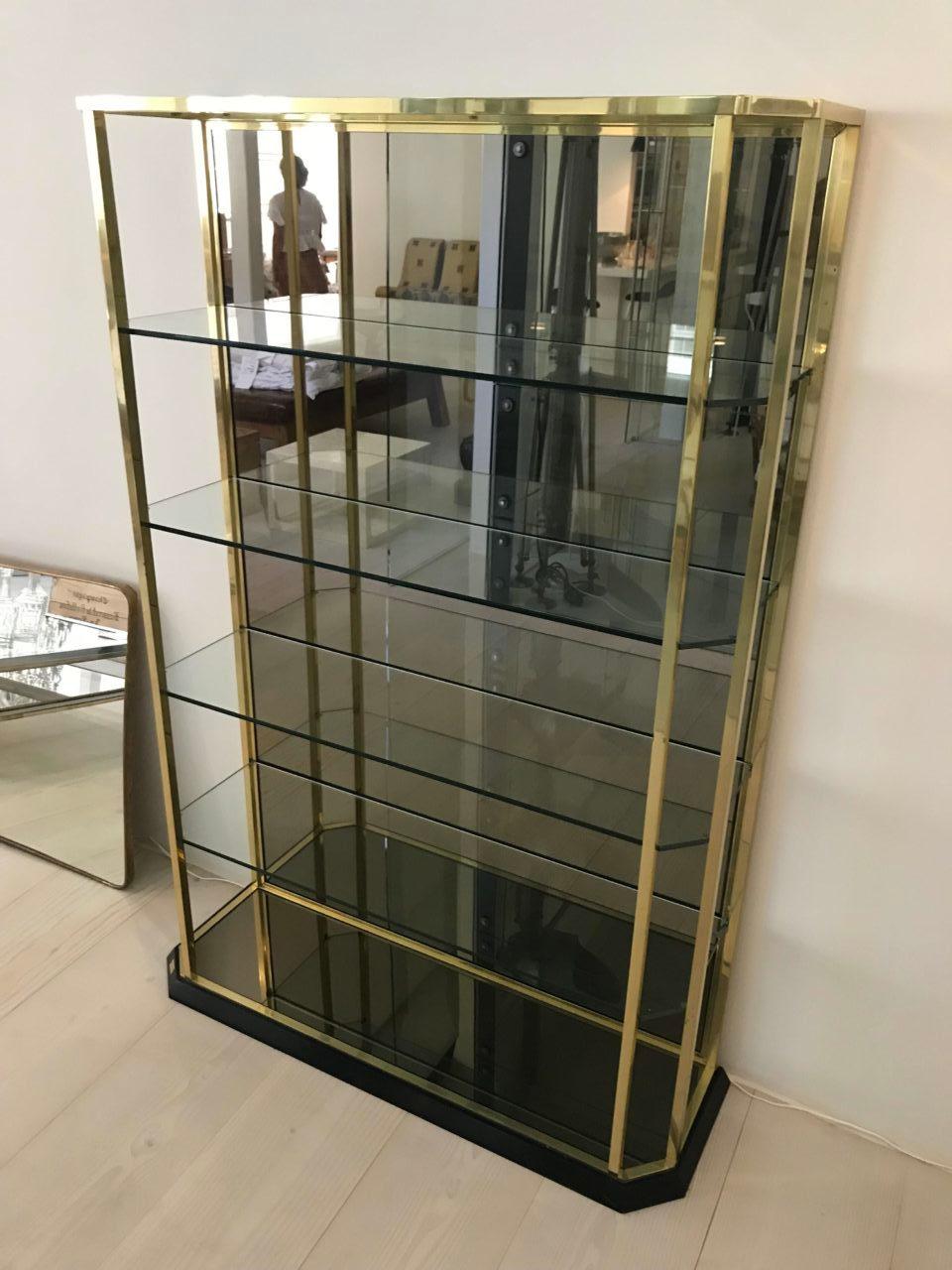 A gorgeous, unique and exclusive Italian shelving unit. Glossily polished brass and mirrored glass. It rests elegantly and seamlessly on a low black lacquered base, has solid quality smoky glass shelves, and the backing is clad in a large mirrored