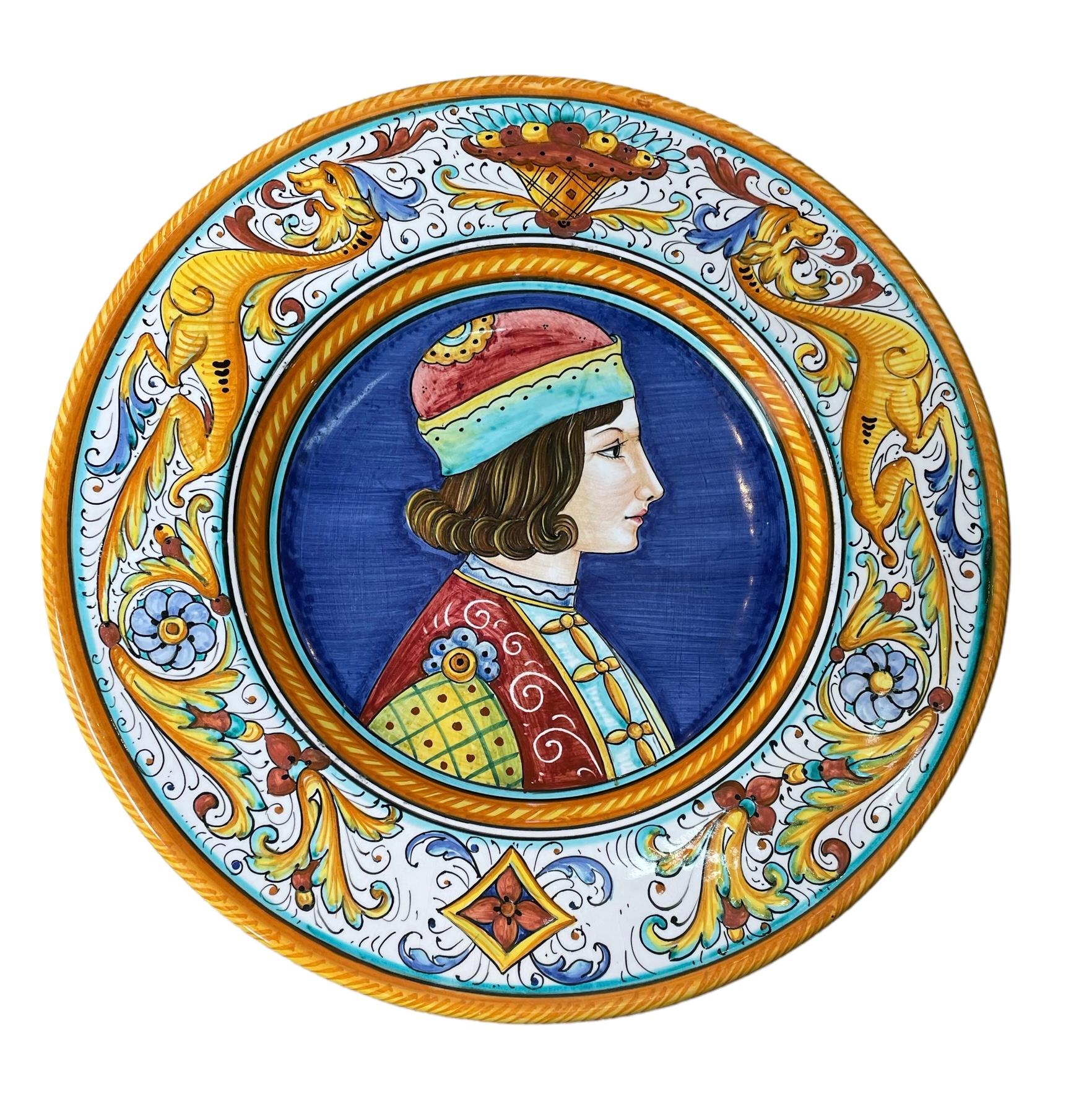 This is a Pair of Gialletti Majolica Plates. It depicts two plates with a portrait of a Renaissance man and lady in the center in a royal blue background. The border of the plate is decorated with mustard color dragons, baskets of fruits, flowers