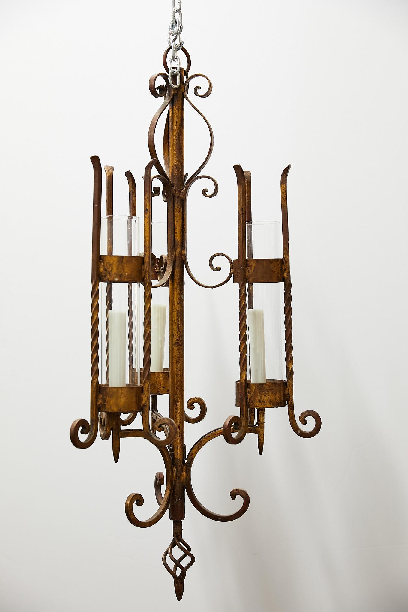 20th century Italian chandelier made of gilded wrought iron having three wired arms with twisted and scrolling candelabras holding waxed candle sleeves enclosed by glass chimney hurricane shades.