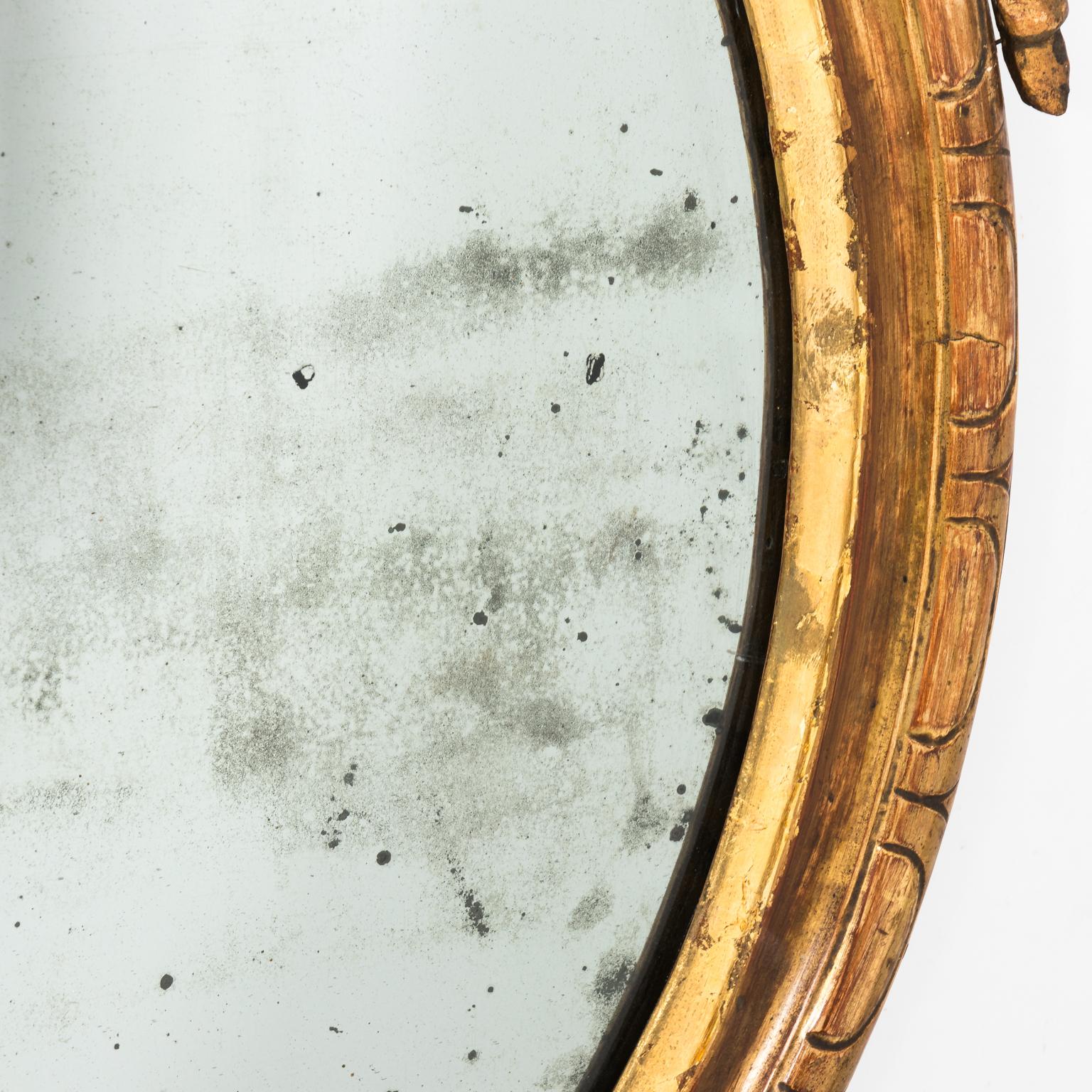 Oval Italian giltwood mirror with carved daffodil on the crown, circa 19th century. Please note the mirror features repair to the crown and chips to the giltwood, the original mirror plate also shows minor losses to silver layer.