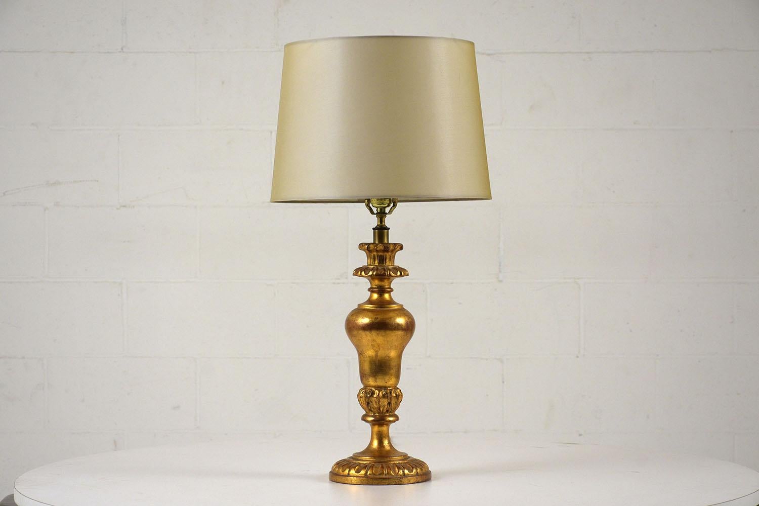 This 1970s Italian Louis XVI-style table lamp features a carved giltwood base with acanthus leaves and egg and dart accents. The lamp is accompanied by a sand color shade in a rayon fabric. This lamp is wired to US standards and is in working