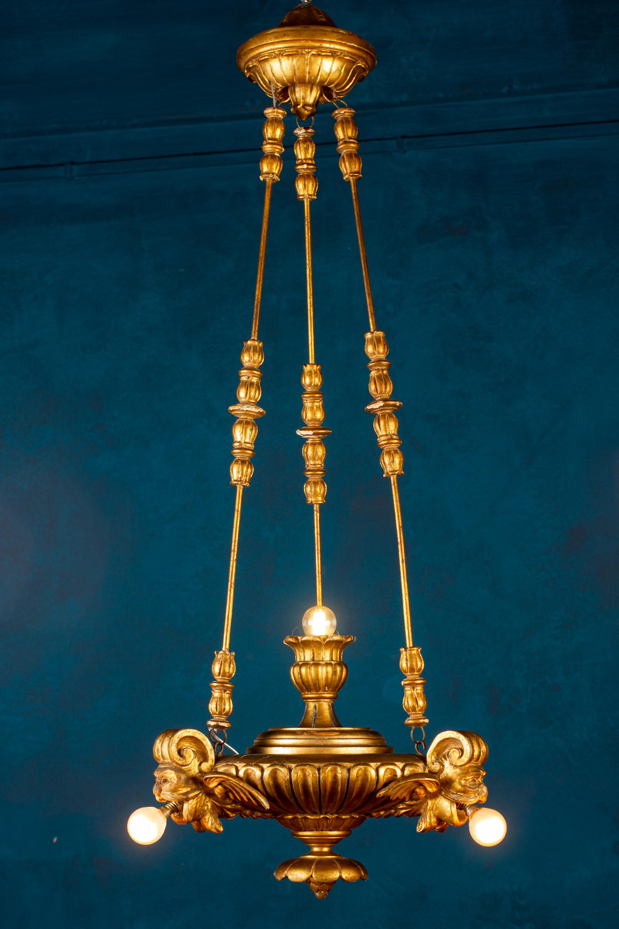 Hochdekorativer, fein geschnitzter und vergoldeter Holzlüster oder Laterne, verziert mit drei geflügelten Drachenmasken und einer Kette, die in einem Baldachin endet.
Vier Glühbirnen E 14.
 