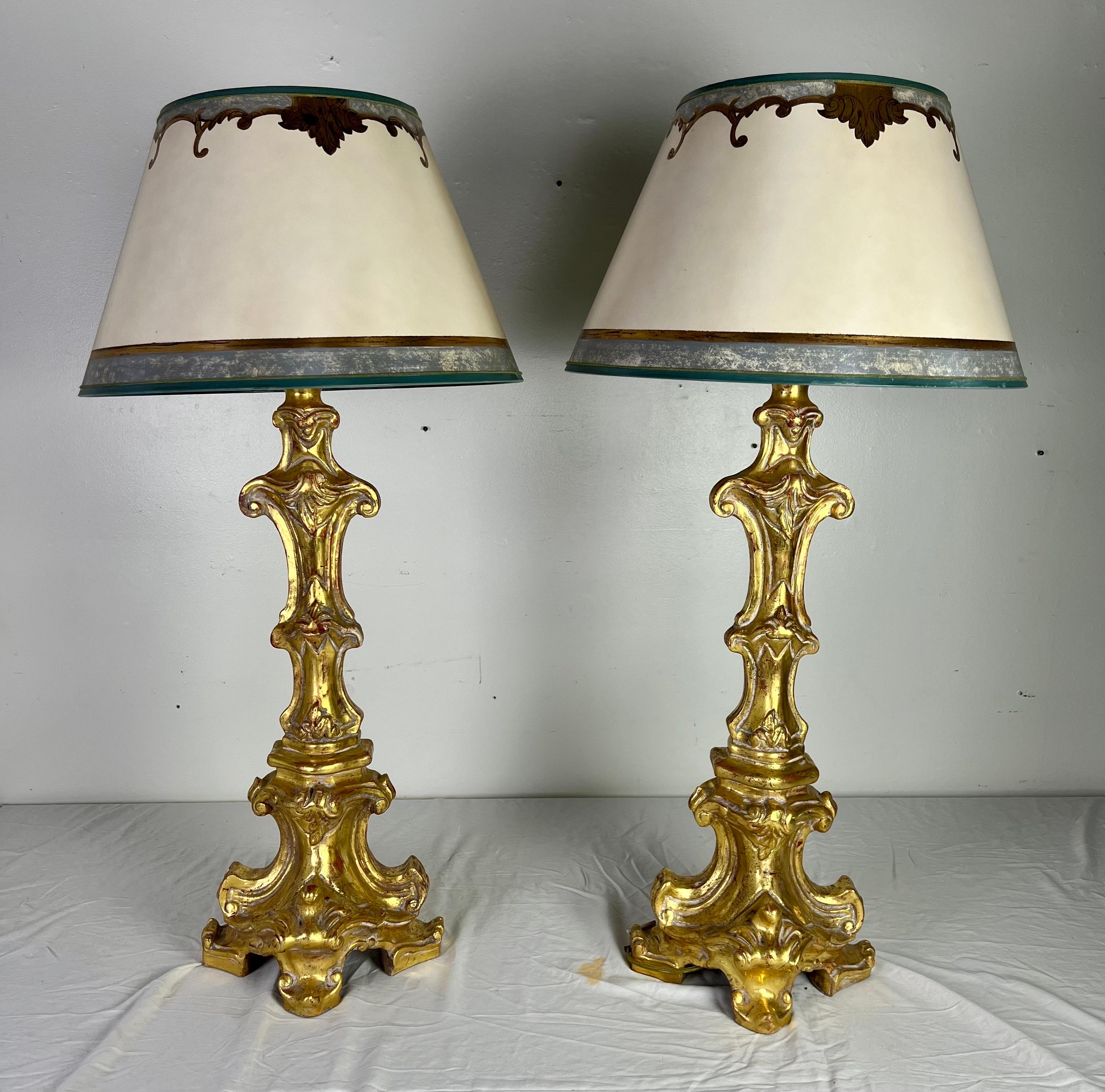 Pair of Italian giltwood candlestick lamps detailed with acanthus leaves and more...The lamps are crowned with hand painted parchment shades in colors of worn blue & gold.  The lamps are newly wired and ready to use.