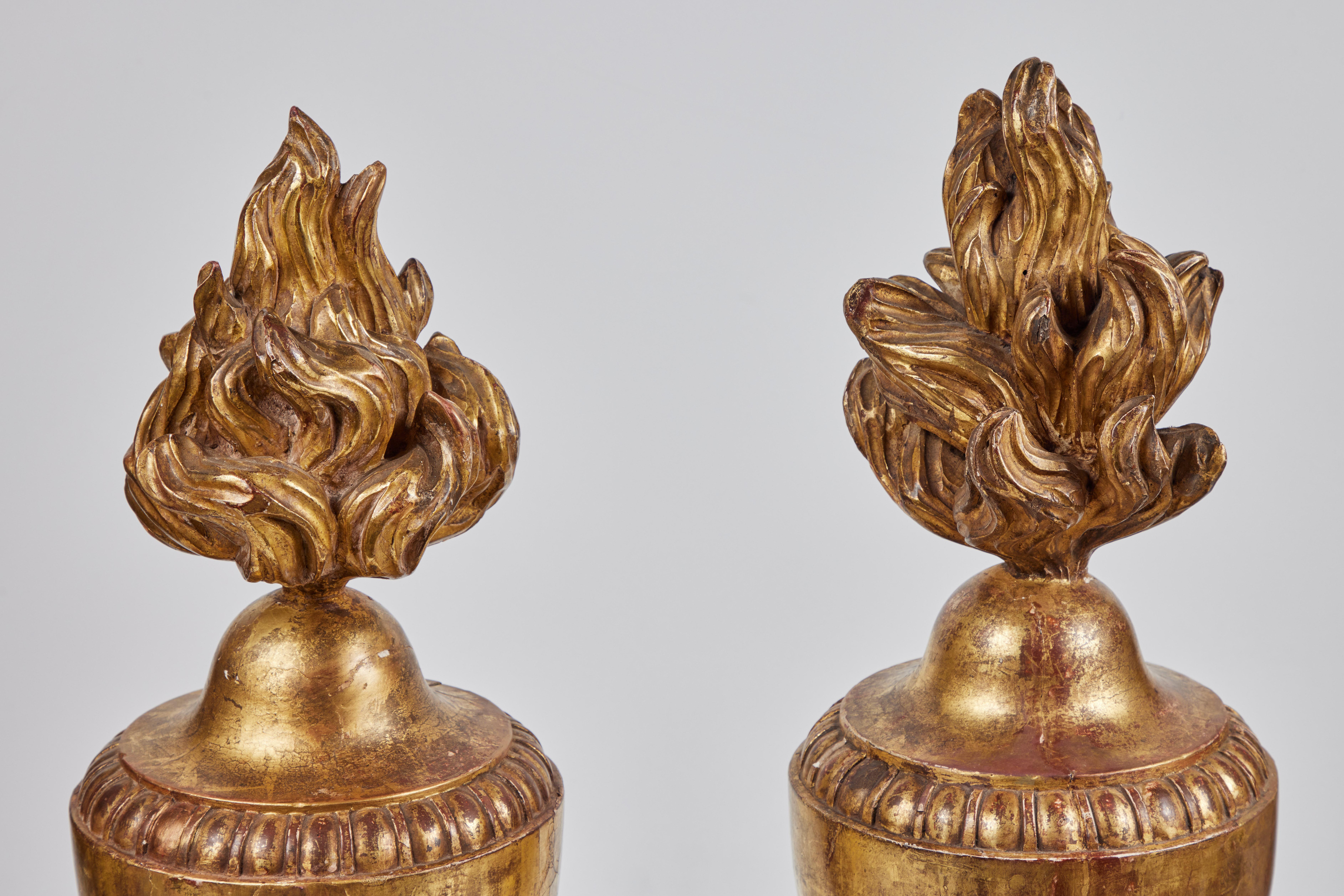 A dramatic pair of hand-carved, gessoed and 22k gold gilded, urn-form balustrade finials surmounted by flame forms. Each on a tapered foot with a pearl relief embellishment.