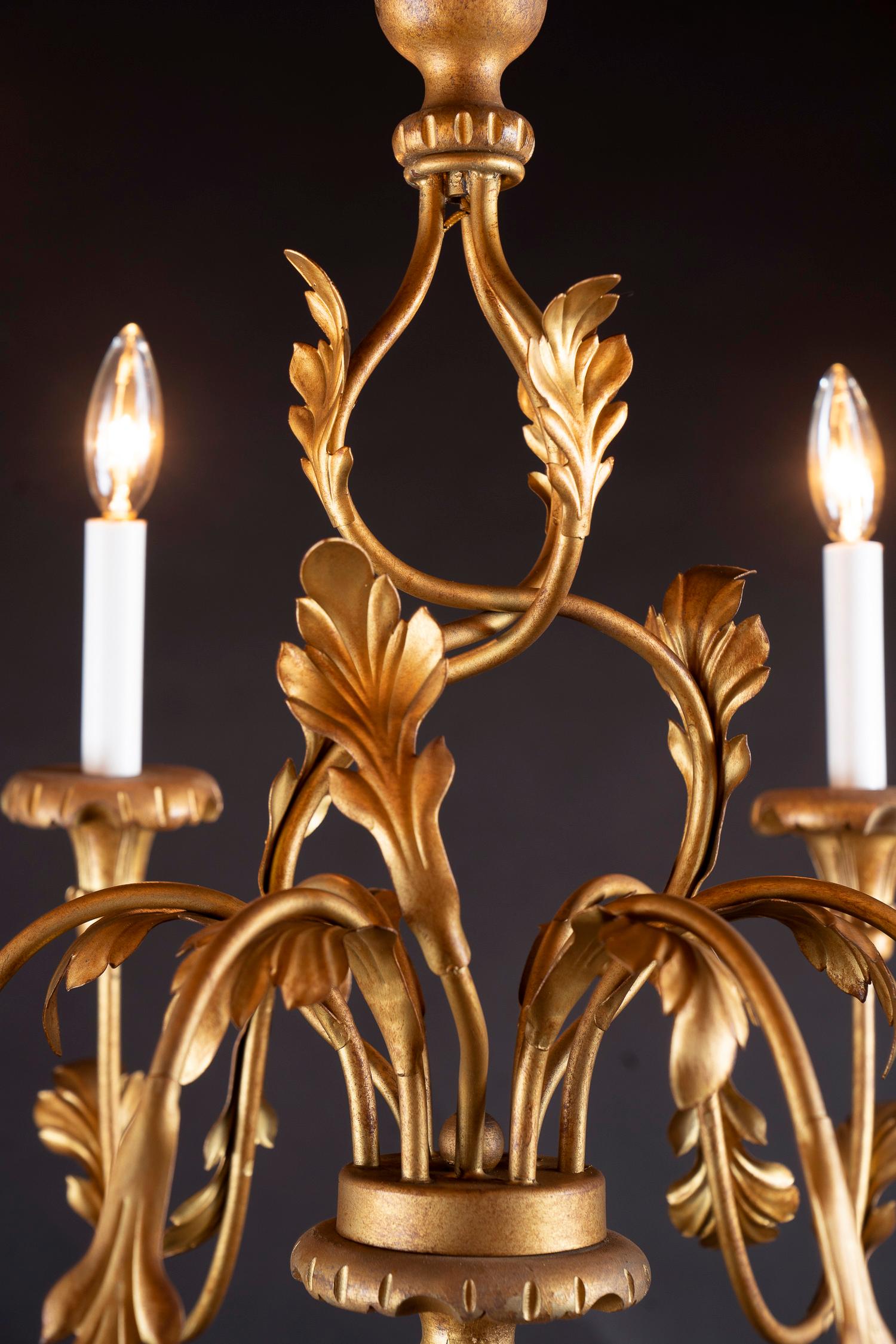 This beautiful Italian chandelier is made of gilded (gold leaf covered) Iron, hand painted wood, and tole. The piece dates back to the mid-20th century and features a lovely spiral aesthetic along its center stem. Decorative furling leaves drape