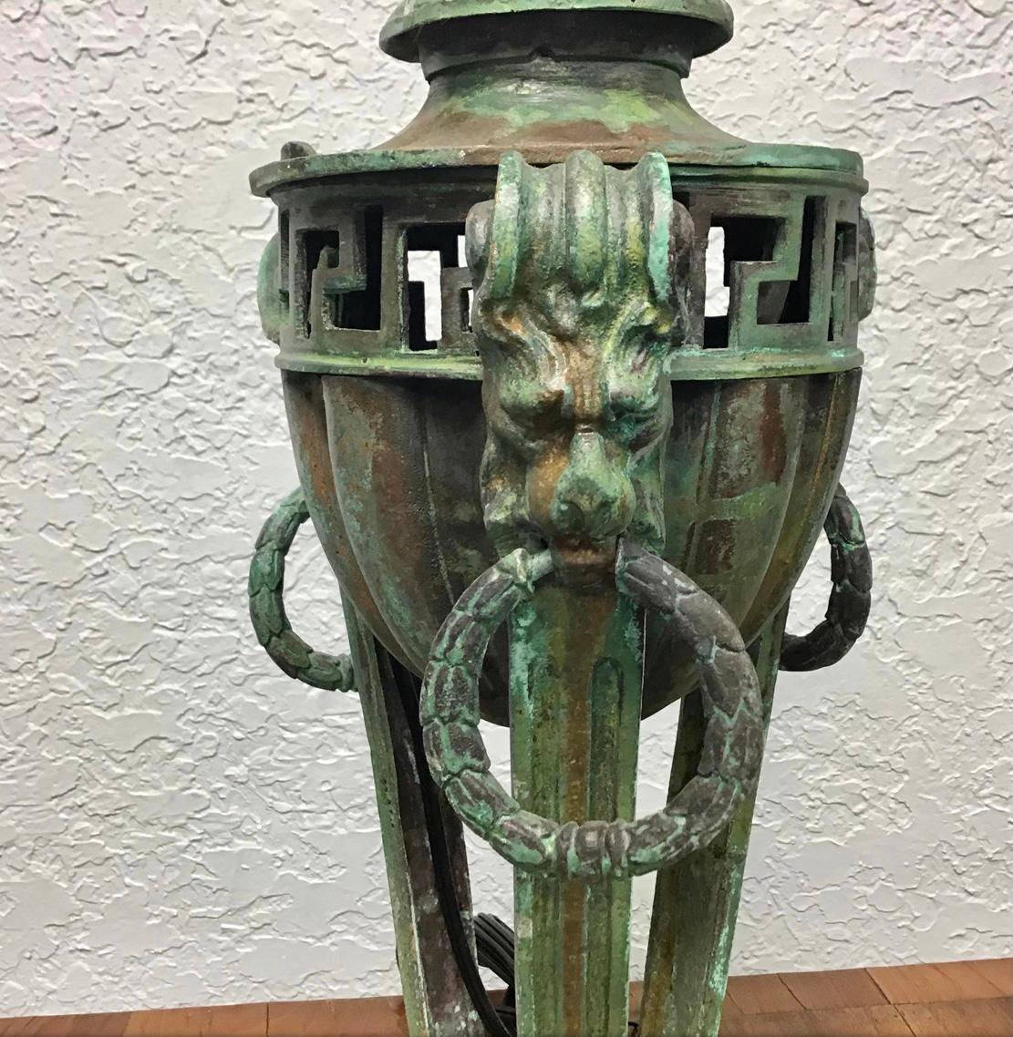 An Italian Grand Tour style patinated metal vessel now mounted as an ancient looking lamp. Paw feet supporting three columns with lion head tops with rings in their mouths. The center in the form of a vessel with open greek key design around the