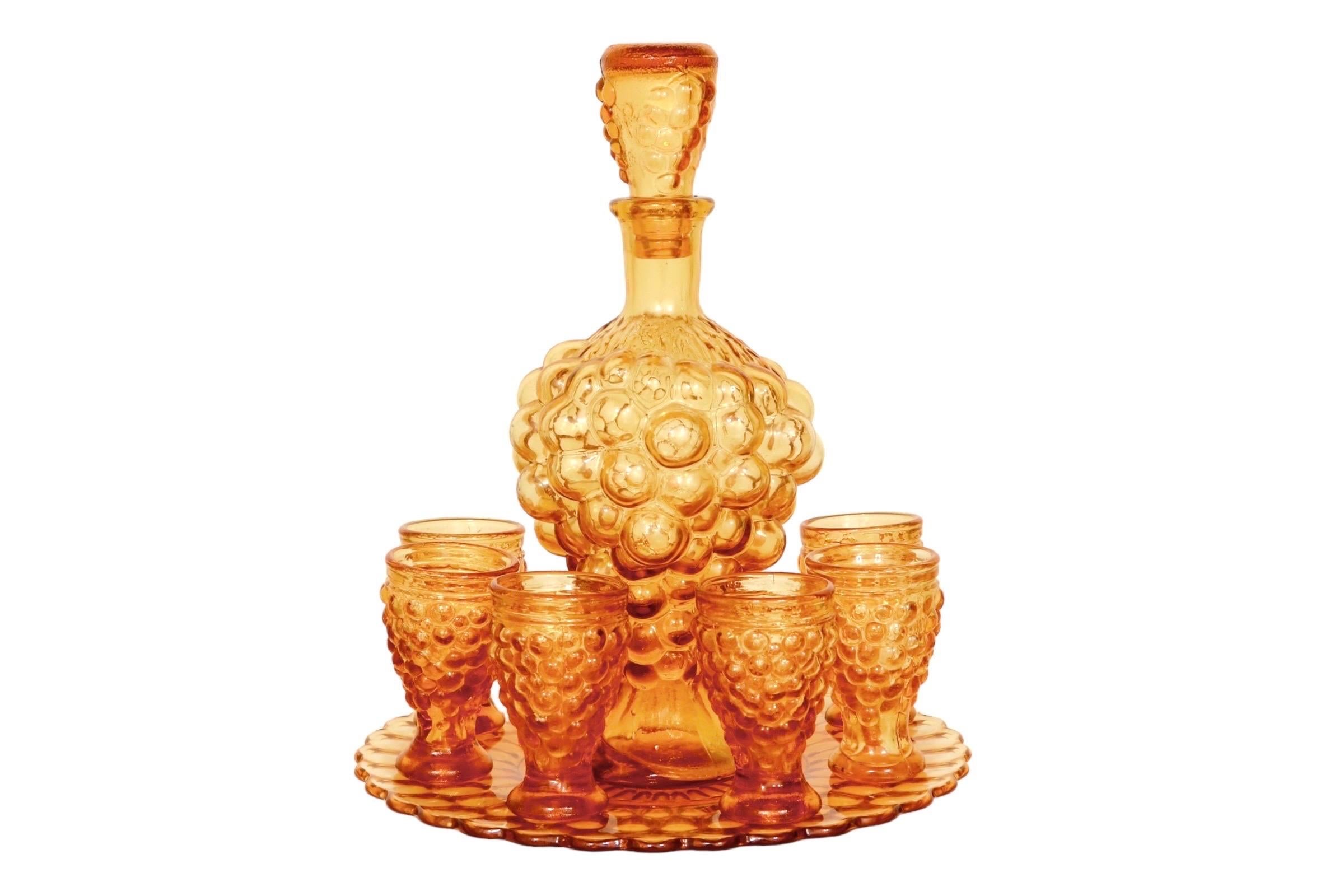 An Italian amber glass decanter set with six glasses and a serving plate. The decanter is pressed in a bubble-like bunch of grapes. The bubble motif is repeated on the decanter stopper, six glasses, and the underside of the plate.