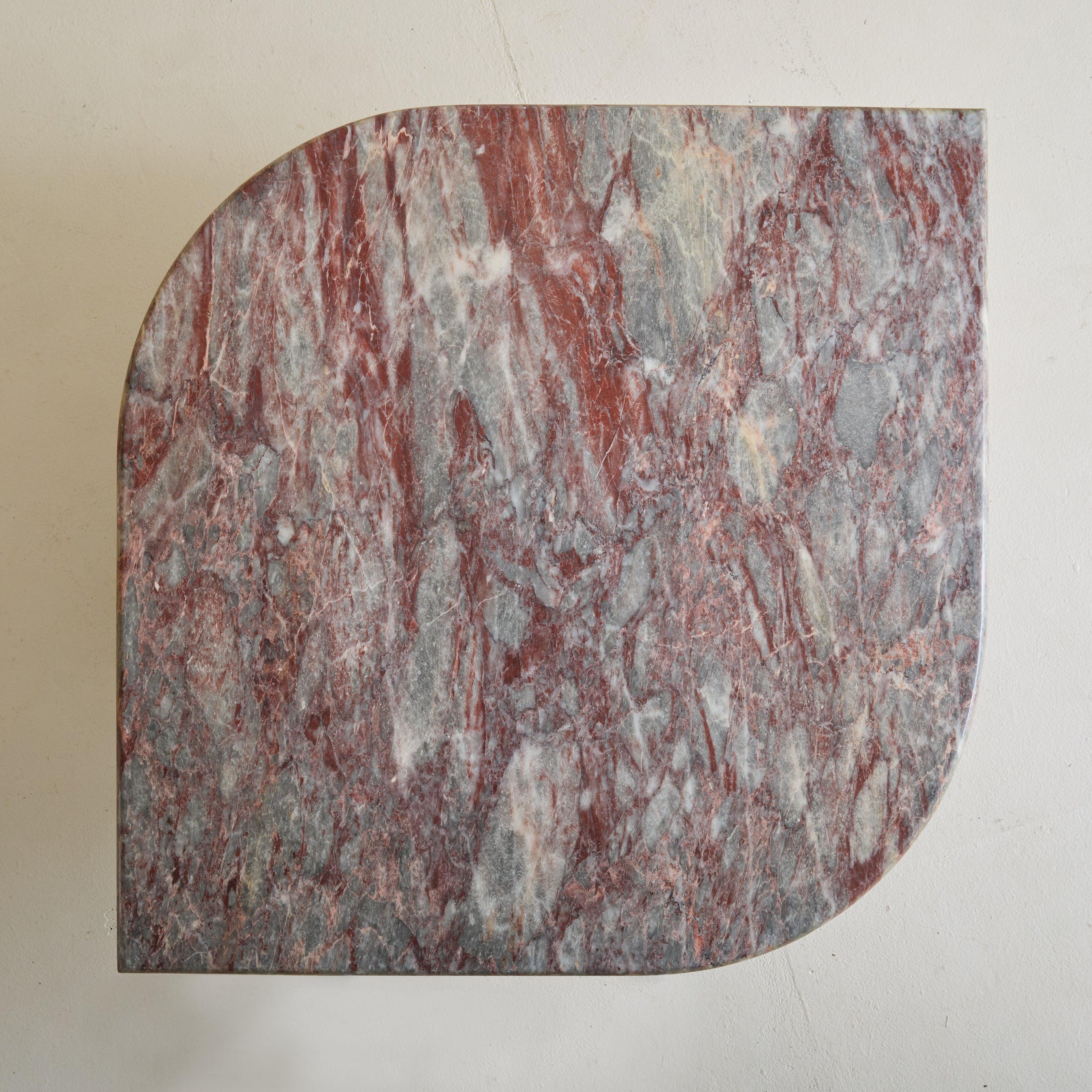 Remarquable table basse italienne des années 1970, fabriquée en marbre adouci dans une variation de teintes grises, lilas et bordeaux. La table présente une dalle ovale pointue qui repose élégamment sur une base à facettes. Probablement issue de