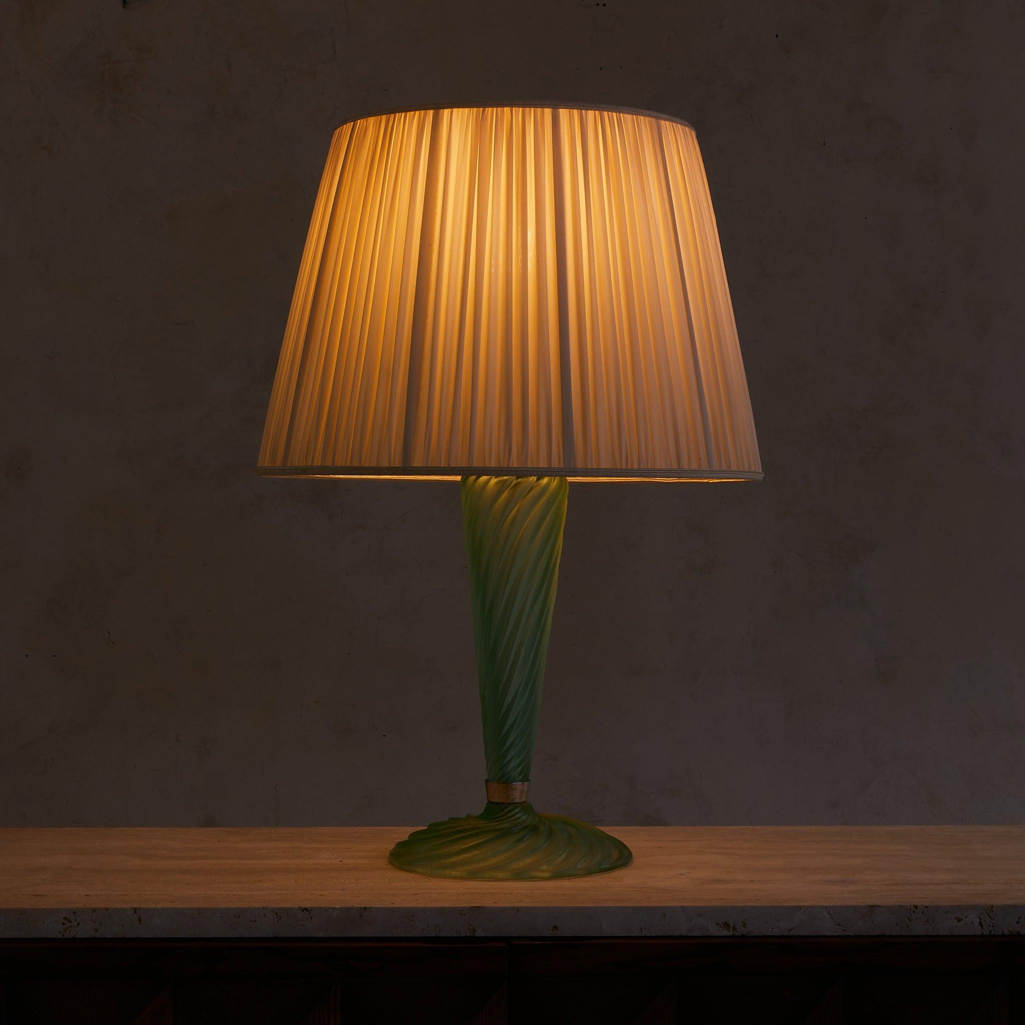 Lámpara de mesa italiana de cristal de Murano, de la década de 1950. La base acanalada está soplada a mano en grueso vidrio verde, trabajado en forma de remolino acanalado y acabado con la técnica del escavado, lo que le da un aspecto suavemente