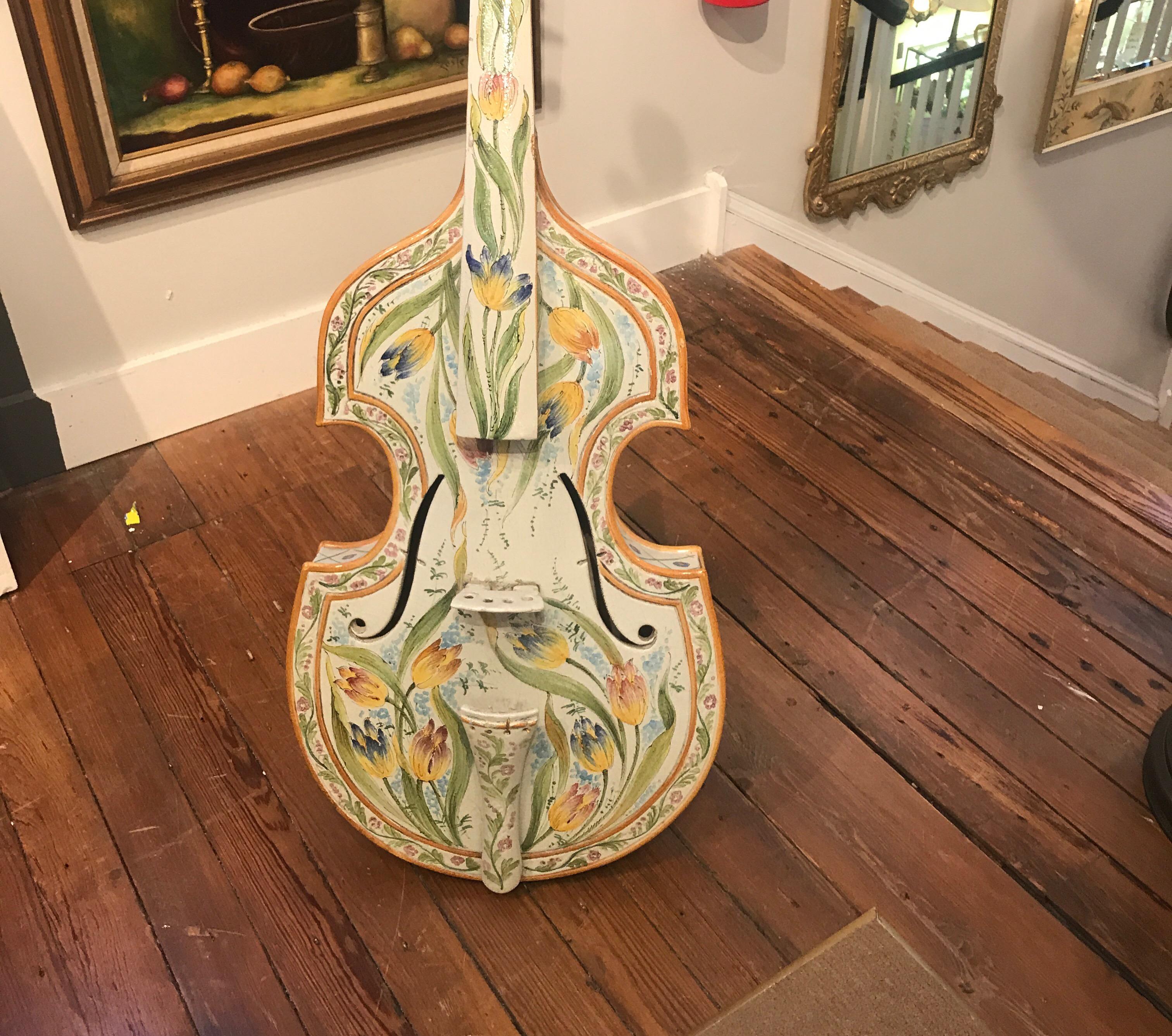 Whimsical hand painted pottery sculpture of a cello. The faience glaze with hand painted floral decoration in vibrant yellow, green and blue, constructed in two parts, the neck and base and fired together. There is a metal sand attached to the