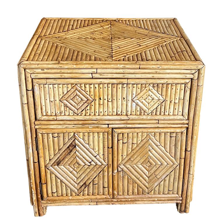 A pair of Hollywood Regency split reed bamboo nightstands or side tables. Strips of split reed bamboo are carefully arranged into a geometric diamond pattern on each side as well as the tops and backs. Each table has a drawer at the top, and doors