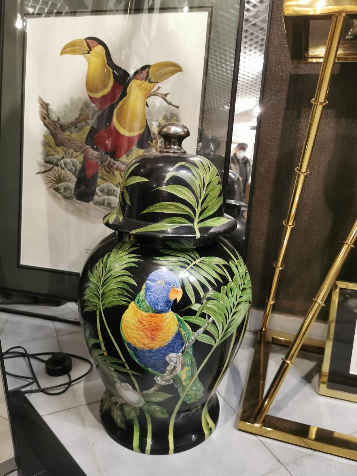 Colourful and exotic artisanal hand painted vase with lid portraiting a blue and orange parrot and vegetation on black background. 
This vase is entirely handpainted by the ceramists of the artisan company 