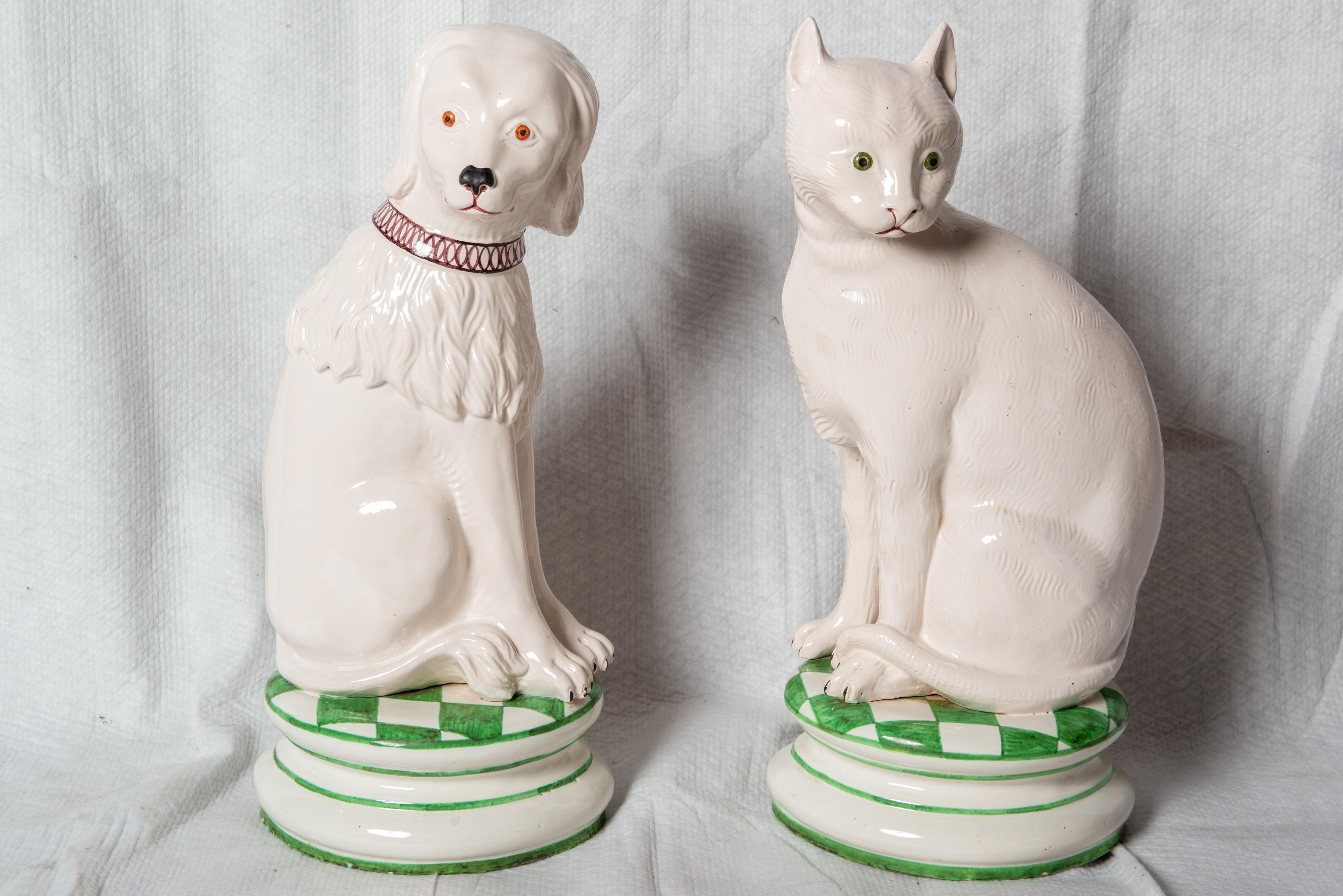 Whimsical ceramic dog and cat statues perched on green and white checked pillows. They are priced individually. Both animals have white fur. The dog has long wavy fur around his neck and shoulders and an elaborate
collar. He has bright brown eyes.