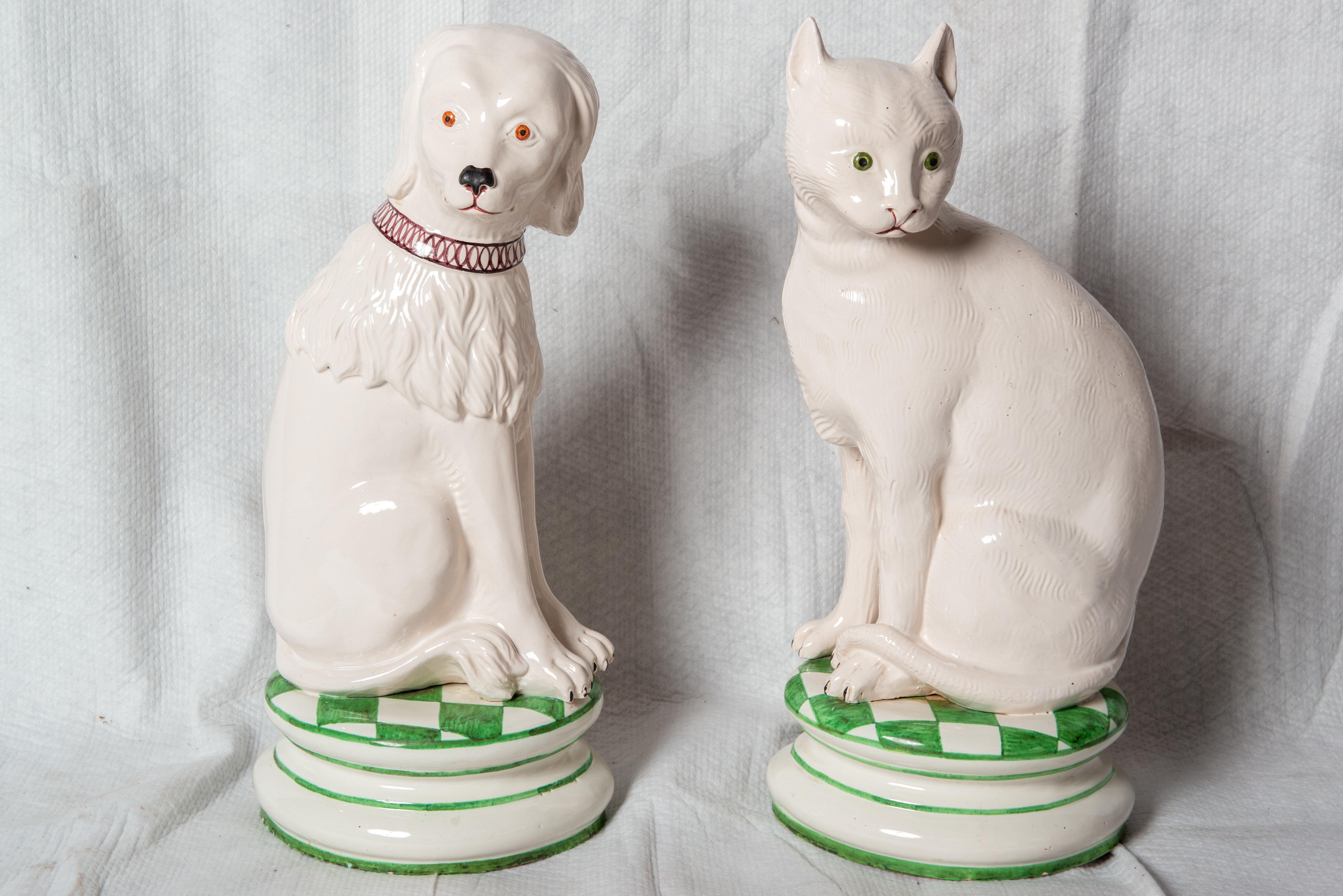 Caprichosas estatuas de cerámica de perro y gato posadas sobre cojines de cuadros verdes y blancos. Tienen un precio individual. Ambos animales tienen el pelaje blanco. El perro tiene un largo pelaje ondulado alrededor del cuello y los hombros y un