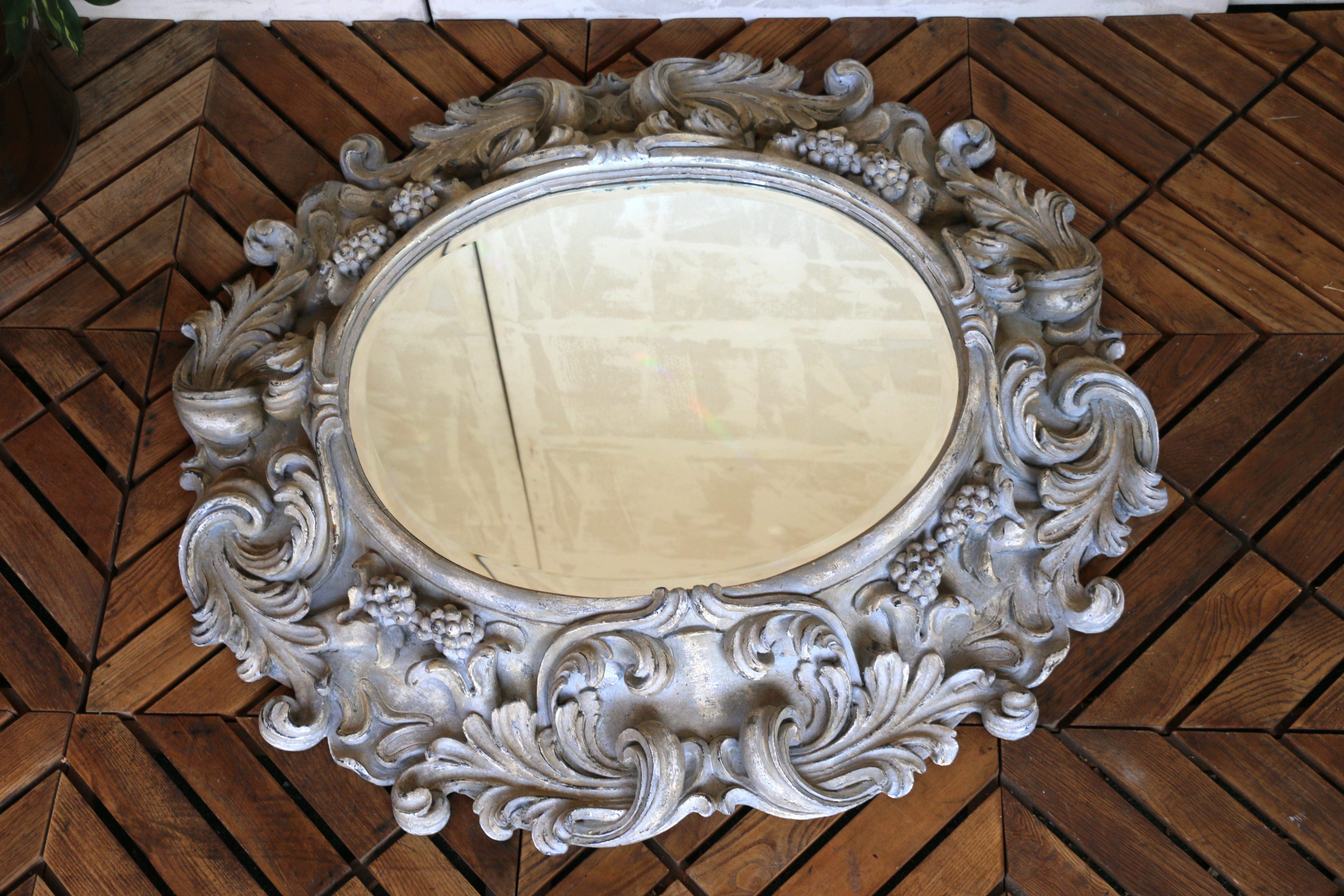 A stunning example of Italian craftsmanship, this large oval mirror is framed by an intricately hand-carved wooden surround, showcasing elaborate scrolling acanthus leaves and clusters of grapes in high relief. The richly detailed carvings create a
