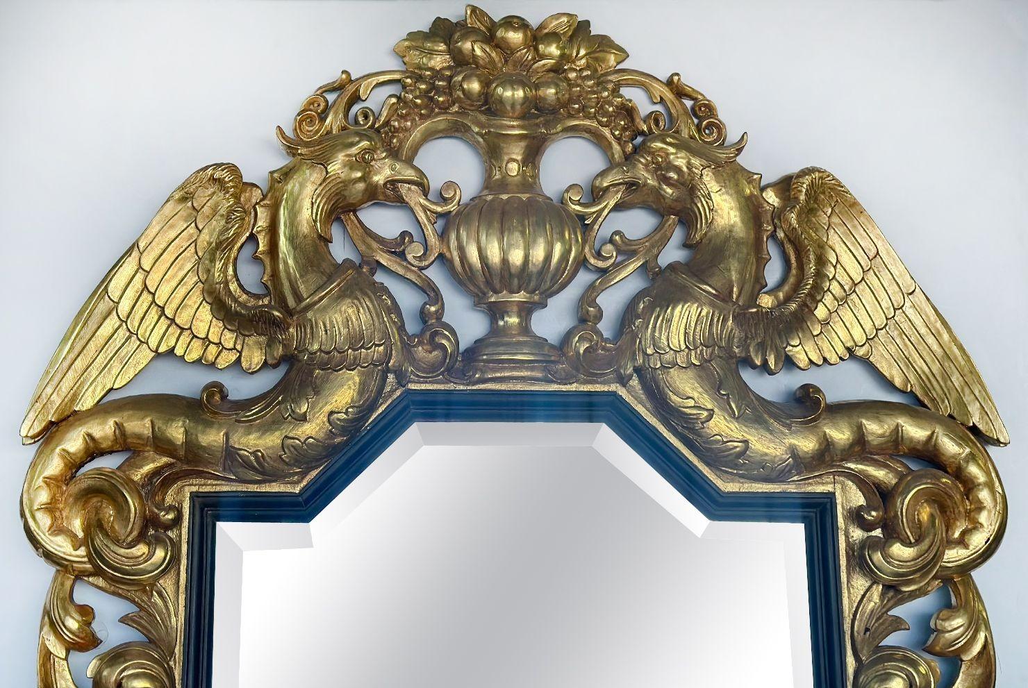 Palatial hand-carved giltwood mirror featuring a 1.5 inch beveled glass. This exquisite piece boasts intricate carved details, including two majestic phoenix mythical creatures adorning the top, alongside an urn figure brimming with an abundance of