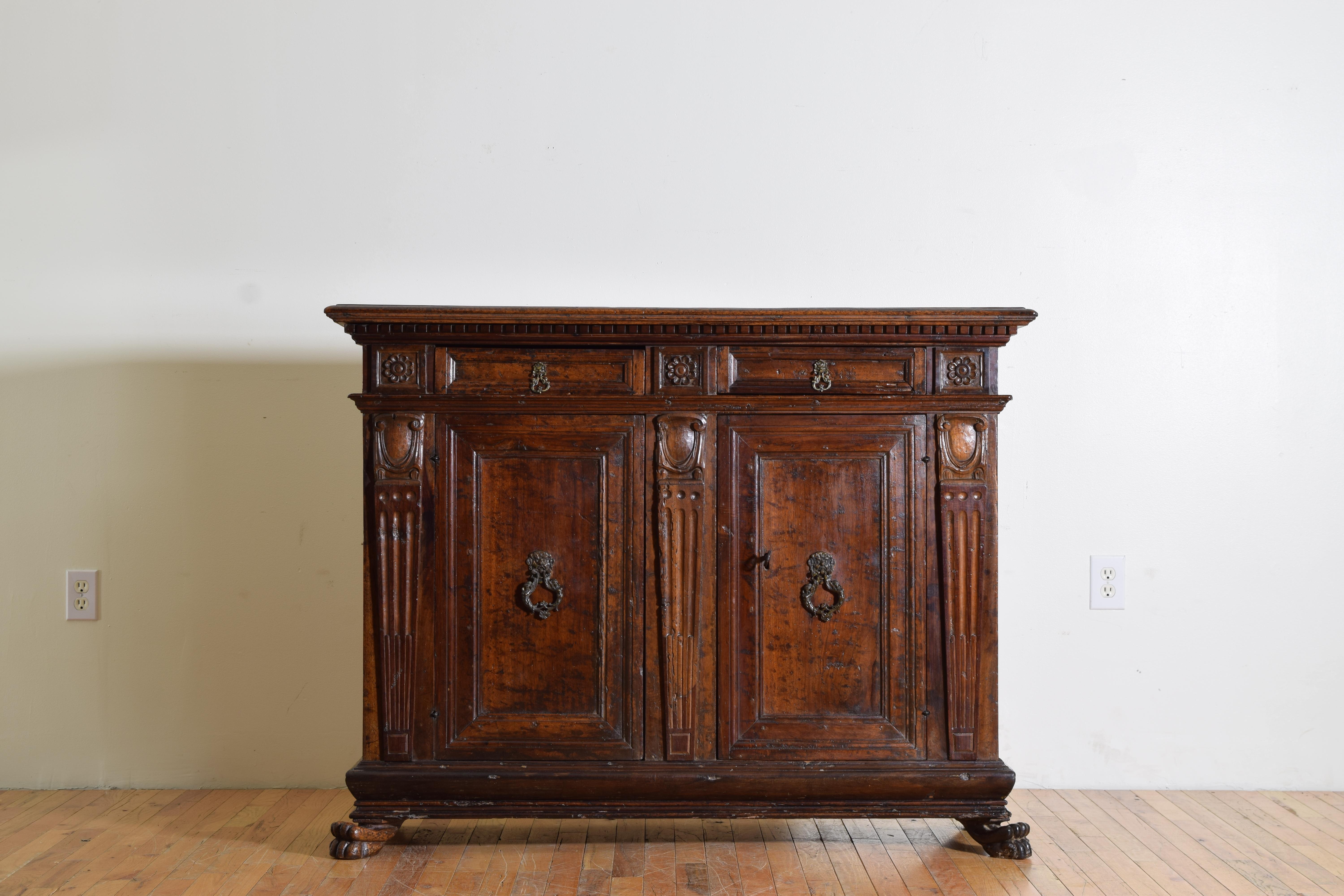 Having a rectangular top with molded and rounded edge with dentile molding below atop a conforming case with two paneled drawers between three framed and carved rosettes, the two paneled doors between three fluted pilasters with carved ornaments