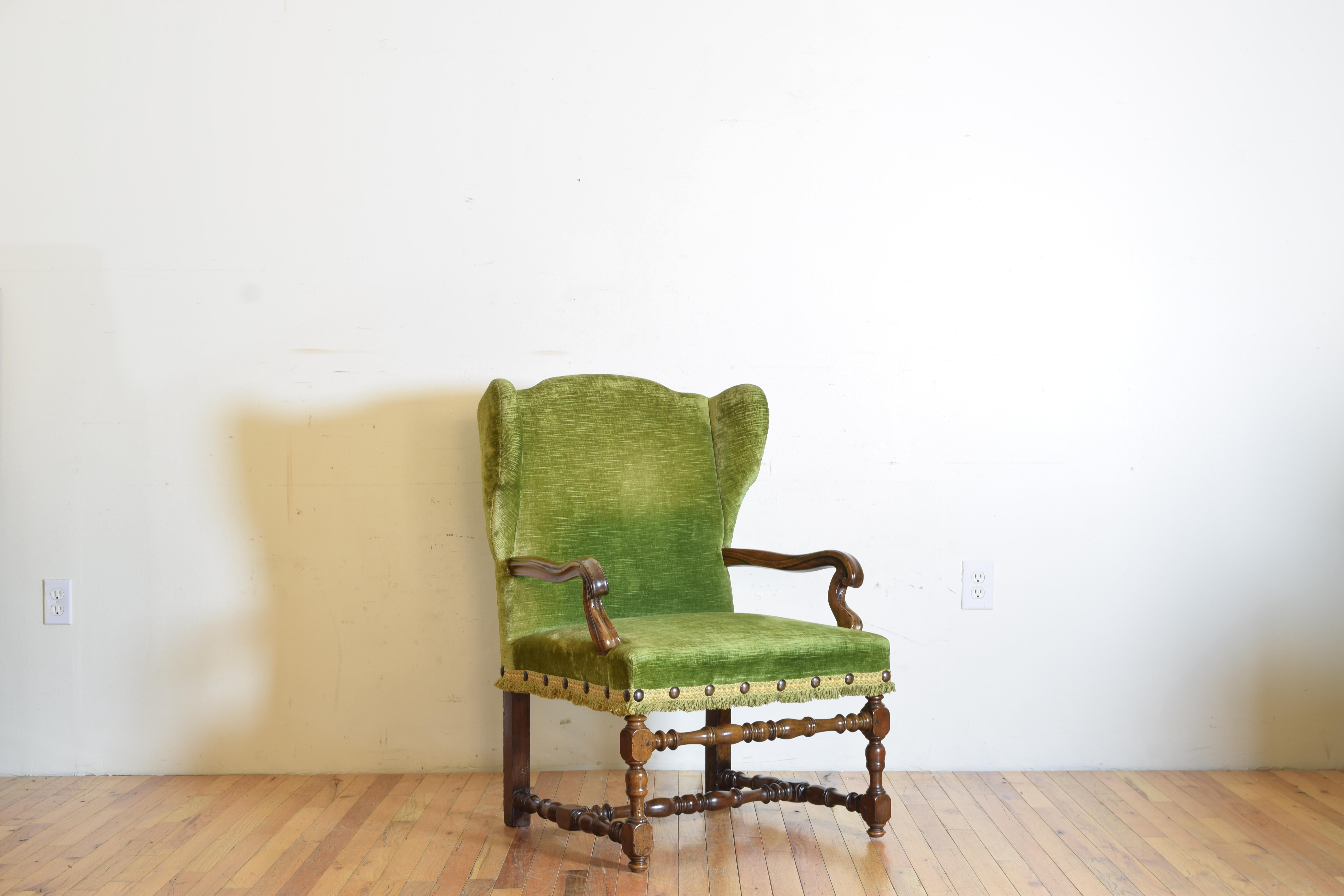 Having a slightly winged back this handsome chair is covered in vintage green velvet with large nailhead anchored tape and fringe, the barely recessed arms above a generous seat raised on turned legs joined by an H-form stretcher, early 18th century.