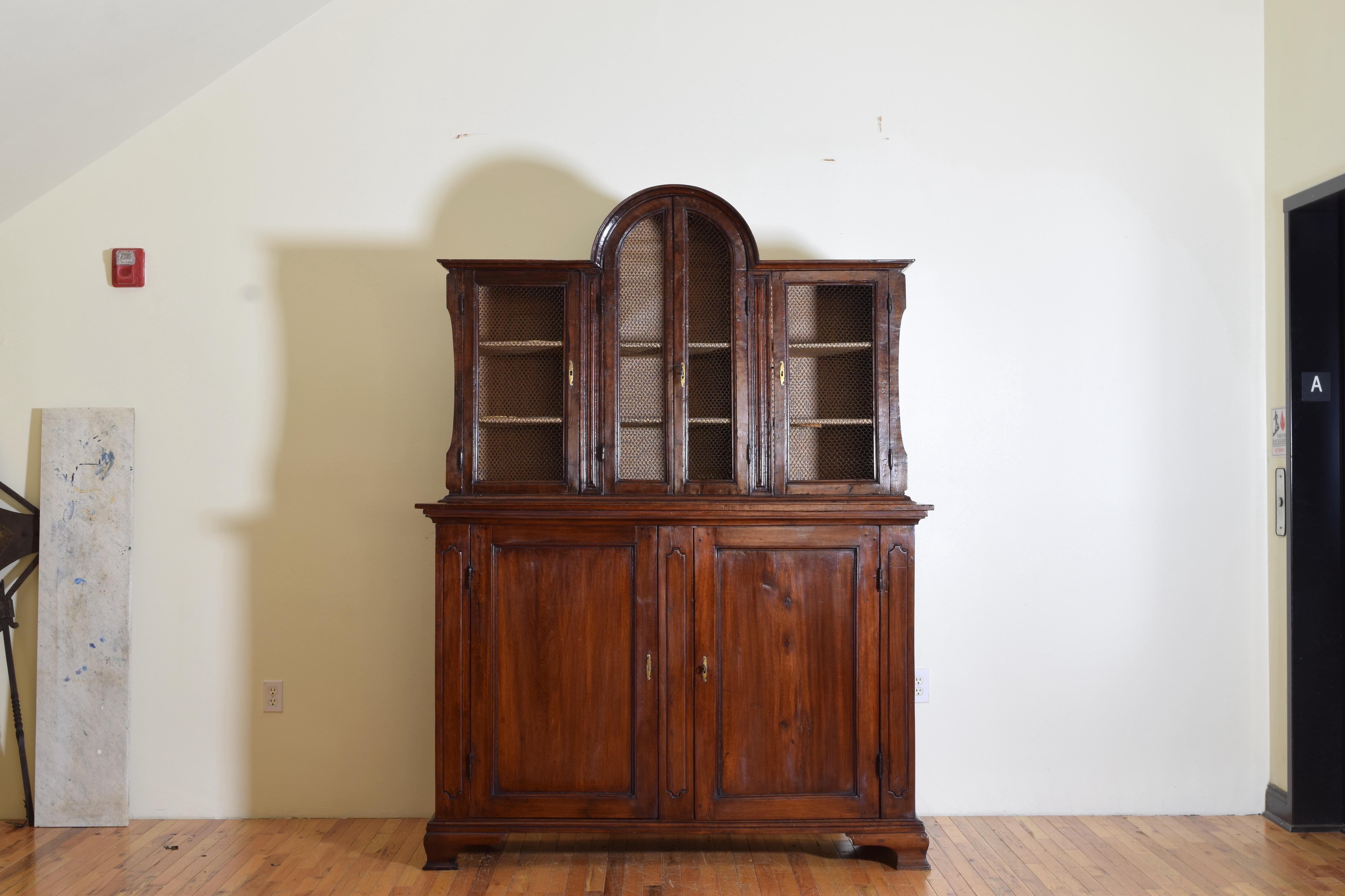 The upper piece with an arched middle section flanked by two equal sections, the locking doors fronted with wire, the lower piece probably of a later date and associated and has two paneled doors opening to reveal interior drawers and is raised on
