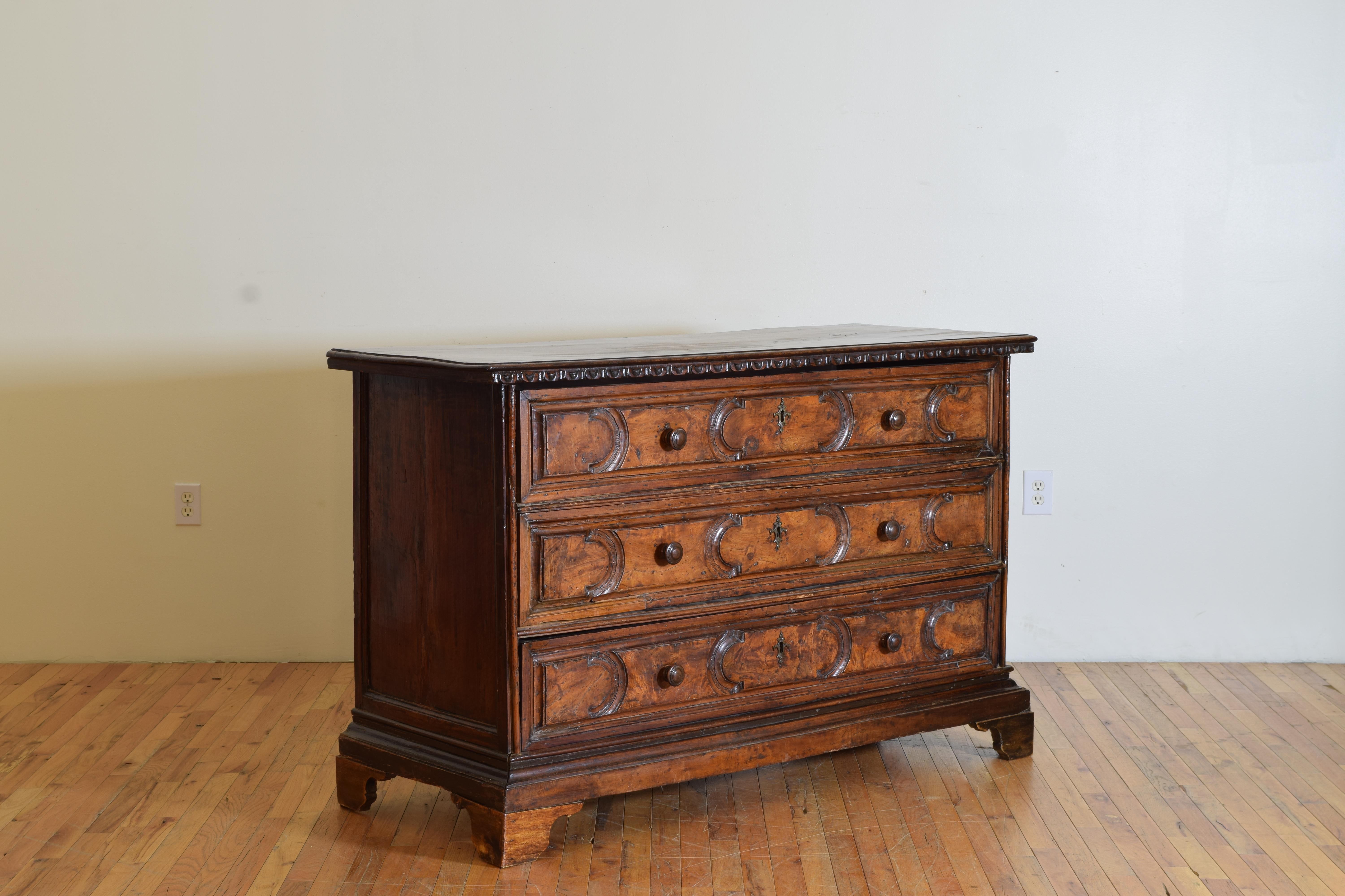 The rectangular top with a molded edge and dentile molding overhanging, the conforming case housing three carved and paneled drawers with original wooden pulls, the sides with raised molded edges, raised on carved bracket feet