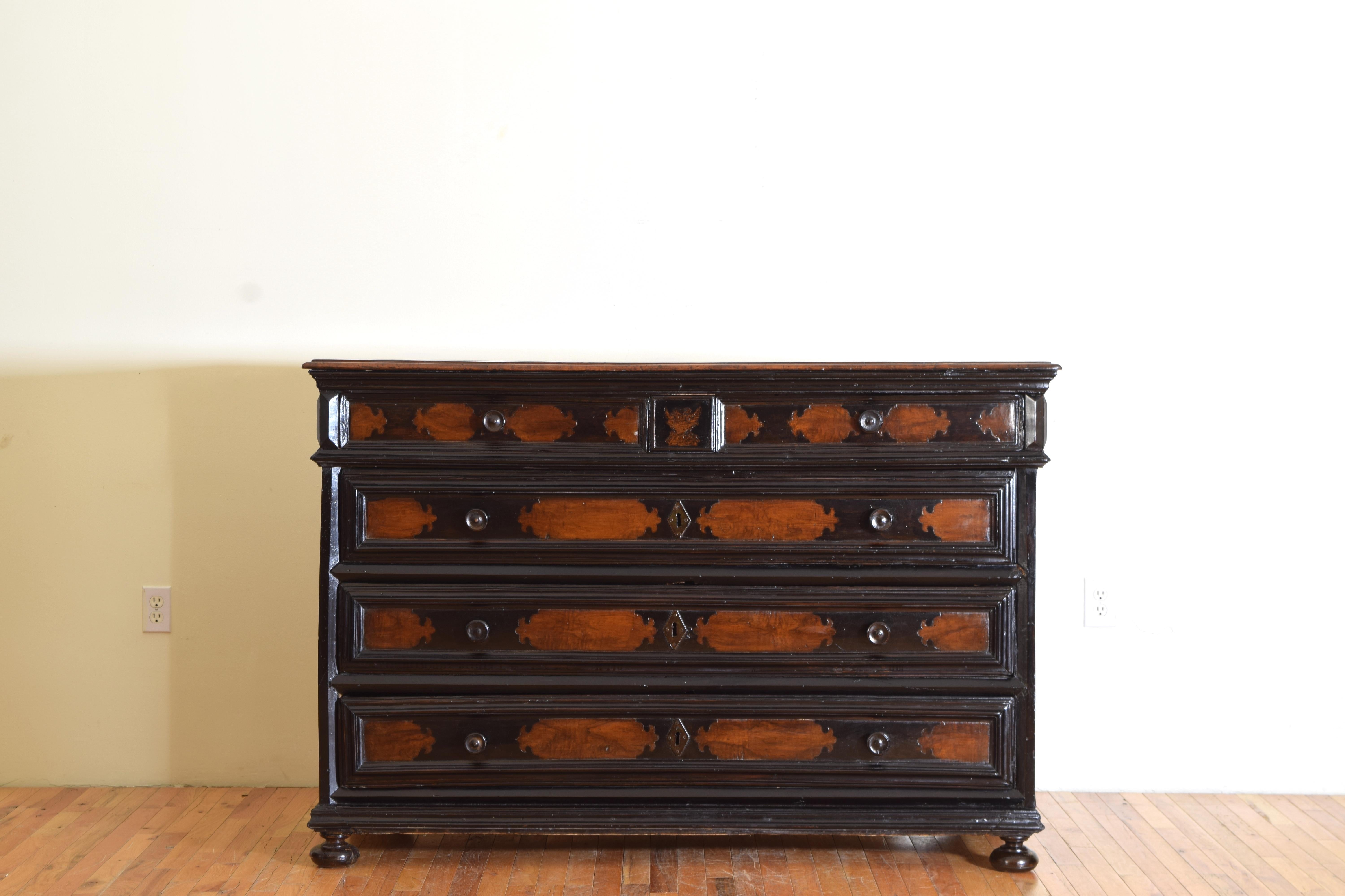 The walnut and partially ebonized body in the rectilinear form resting on ebonized bun feet, three large drawers with an additional two smaller upper drawers, the front panels embellished with patterns of contrasting color.