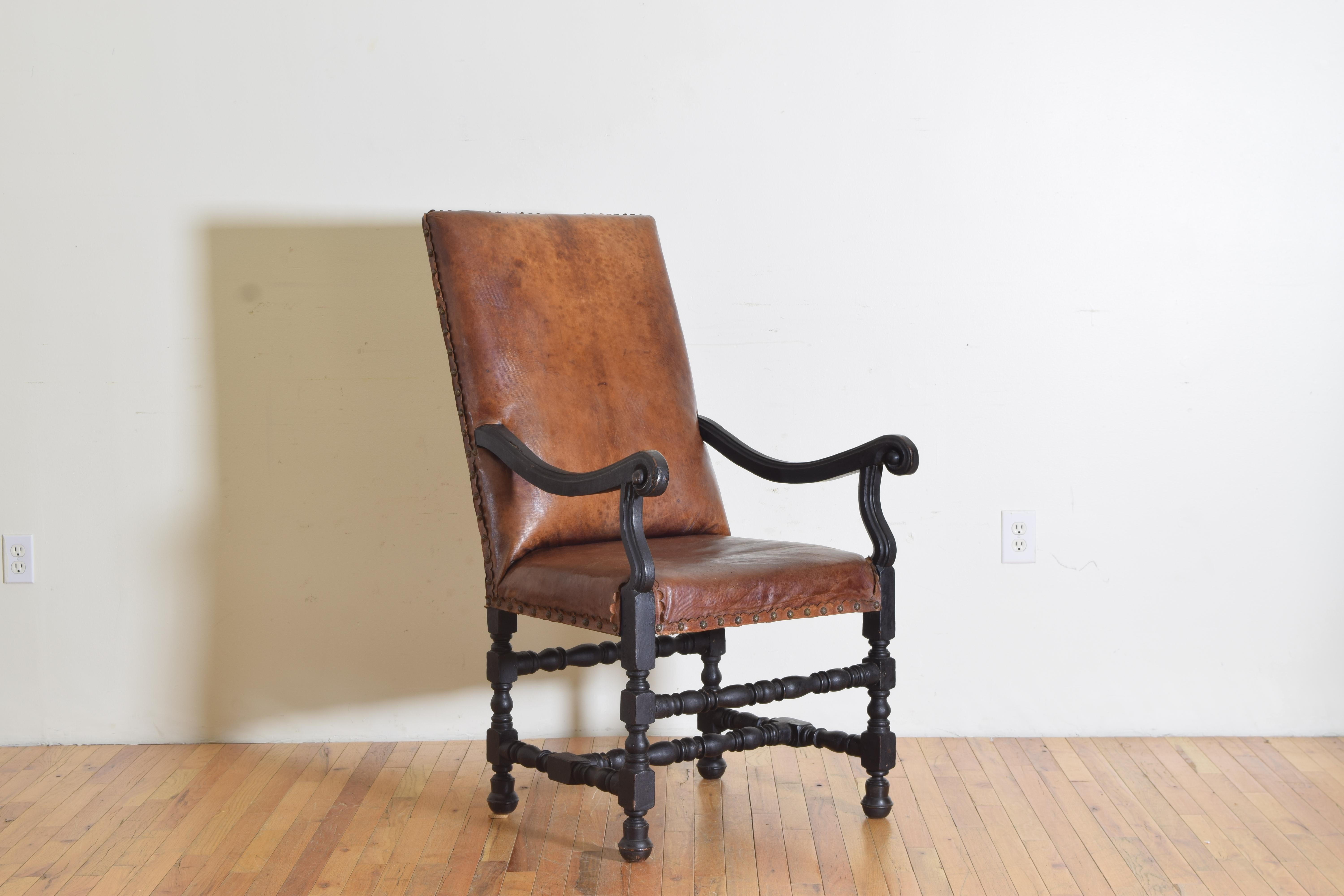 having a slanting rectangular backrest and tight seat upholstered in antique leather and trimmed in cast brass nailheads, the chair retaining its antique ebonized finish, with slightly outwardly scrolling arms raised on scrolled supports, the frame