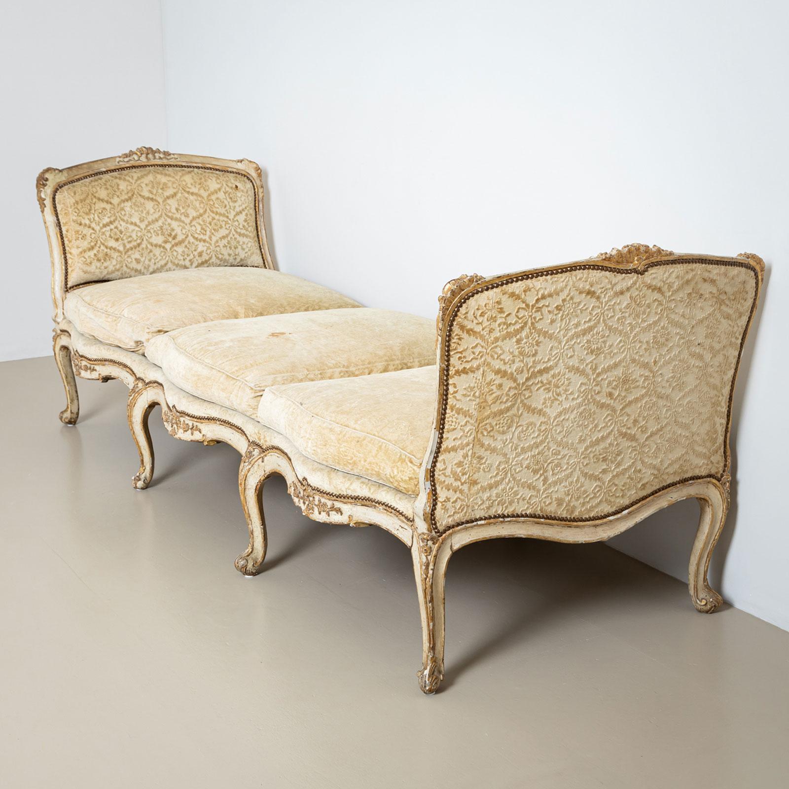 Large Louis Quinze chaise longue with curved legs and intricately carved floral decorations on the apron. The upholstered and rounded side panels add an elegant and inviting touch to the piece. The frame is finished in a white patina with gilded