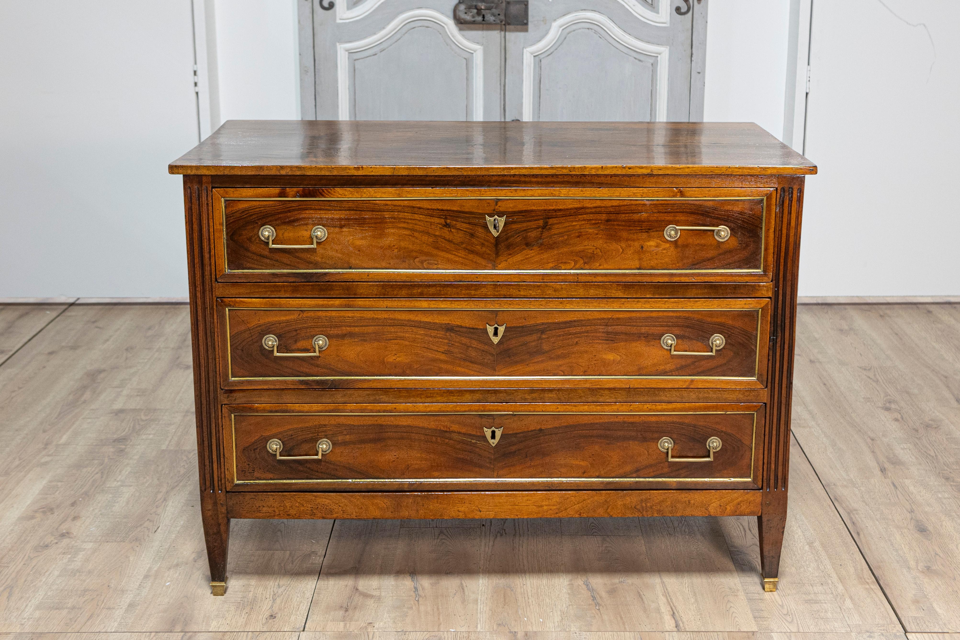 An Italian Louis XVI period walnut commode from the late 18th century, with three drawers, brass trim, fluted side posts and tapered legs. This exquisite Italian Louis XVI period walnut commode from the late 18th century exudes elegance and refined