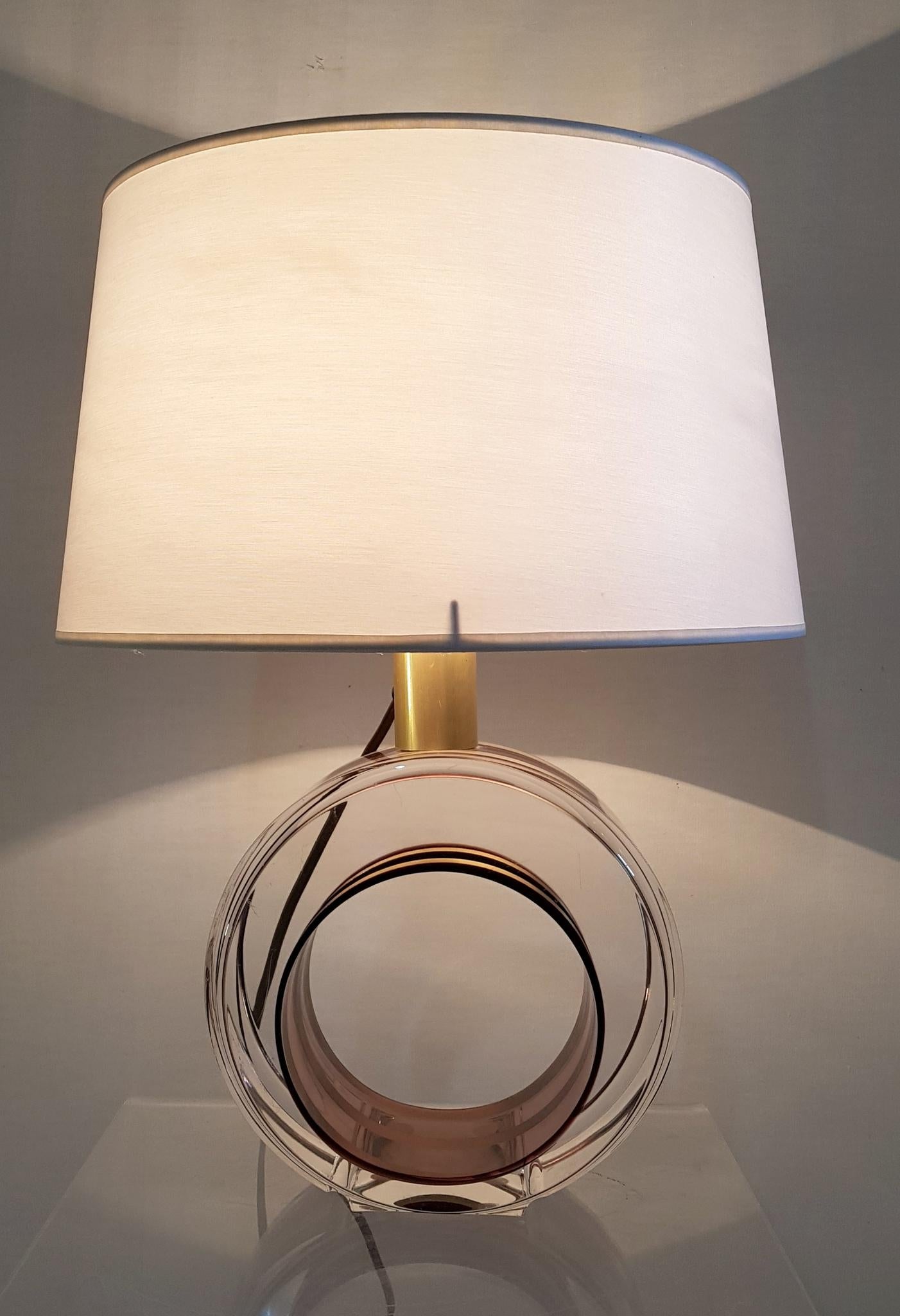 A table lamp with round base in Lucite with a swirl design inside in gold colored aluminium. The color of the base changes a bit with the light but is somewhat purple/amber colored. Comes with a lampshade in white cotton.